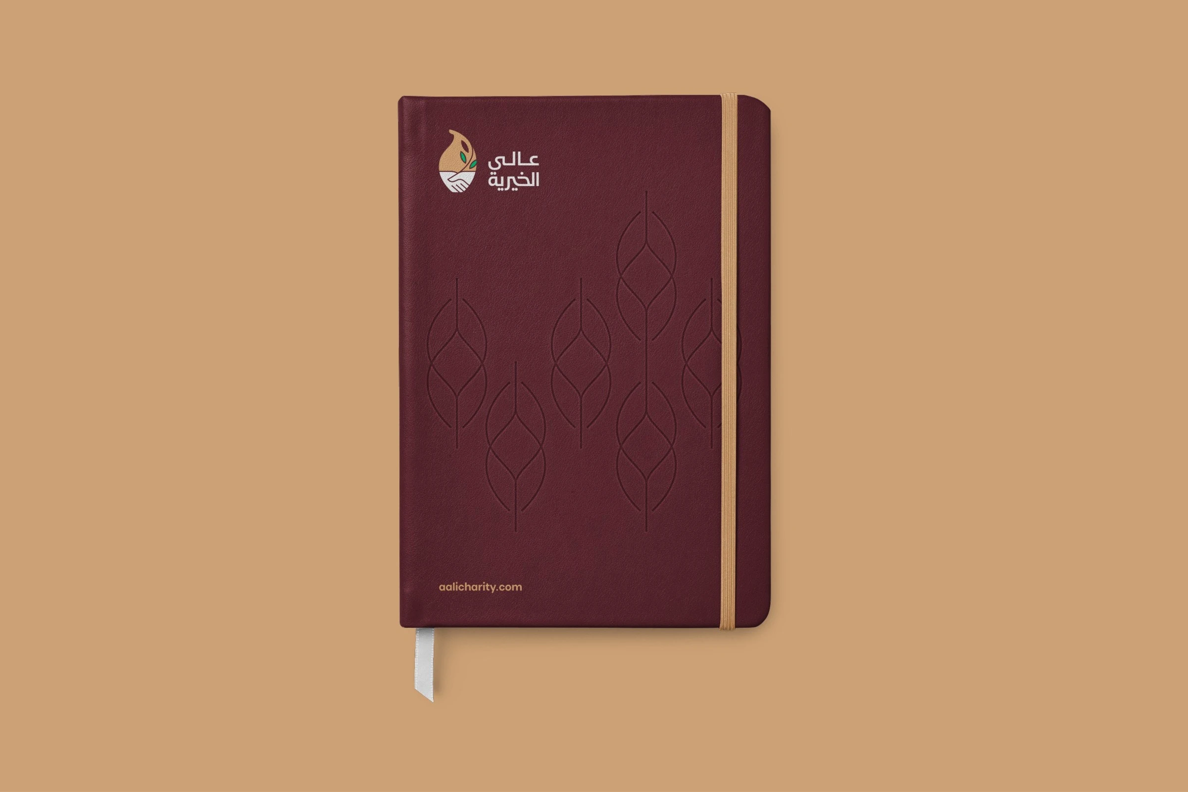 A maroon notebook with the short logo and the leaf motif embossed on its cover, closed by a sand-coloured band.
