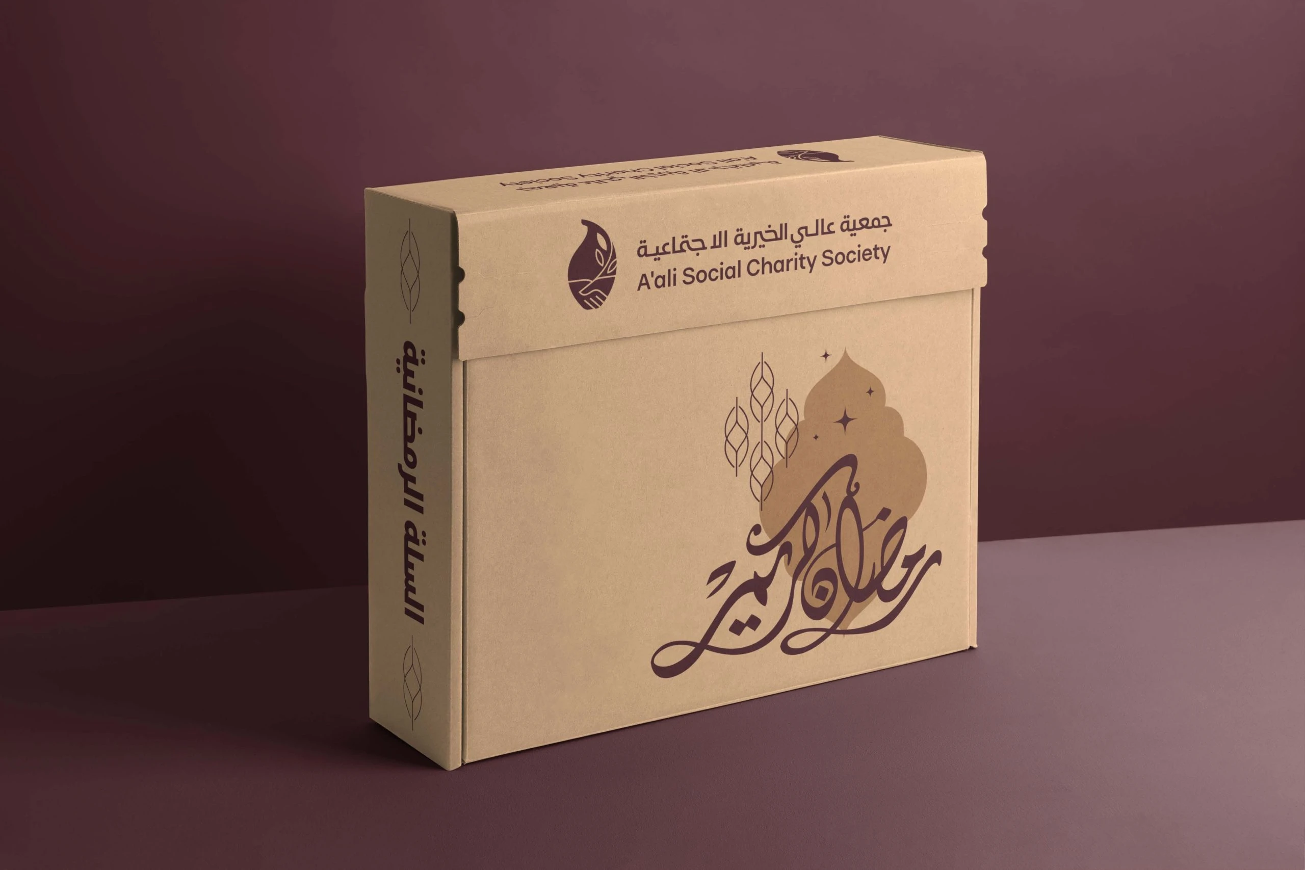 A kraft cardboard box printed in maroon with the logo, Arabic calligraphy and a lantern drawing, for Ramadan basket donations.