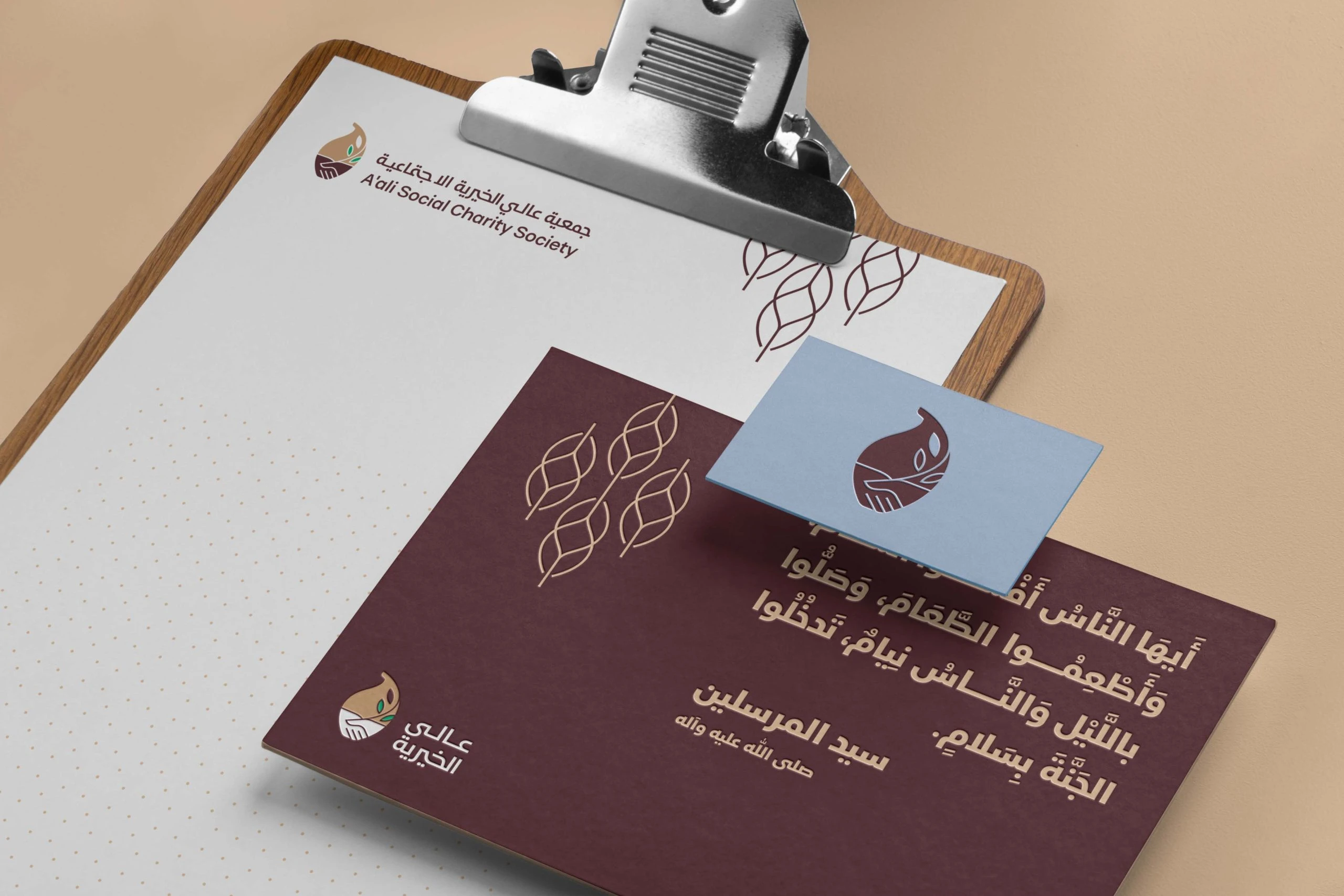 Letterhead on a clipboard, a maroon card with an Arabic saying in sand-coloured type and the leaf motif, and a pale blue card with the emblem.