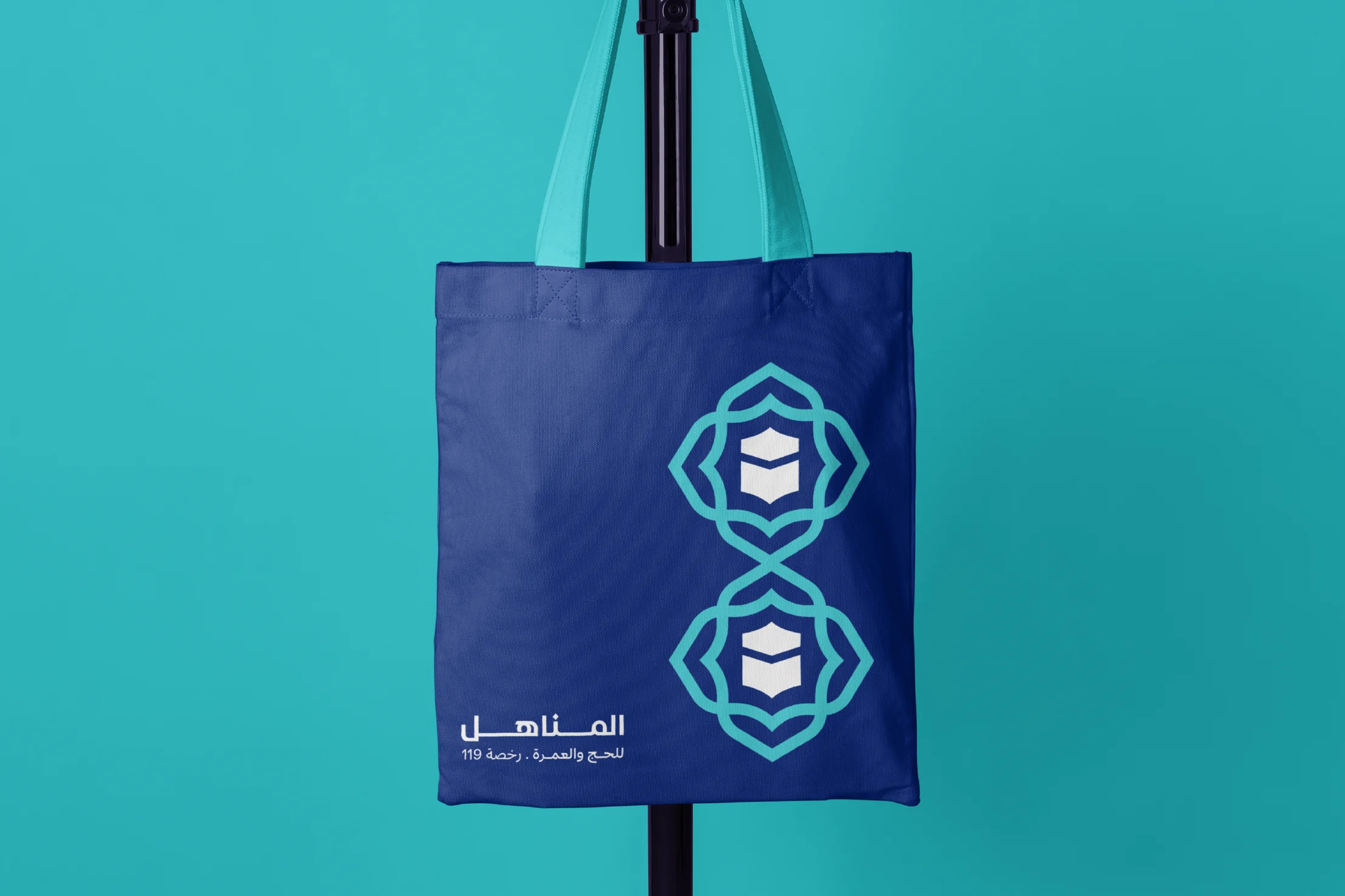 A navy tote bag with two emblems one above the other and the Arabic logo, hanging on a stand against teal.