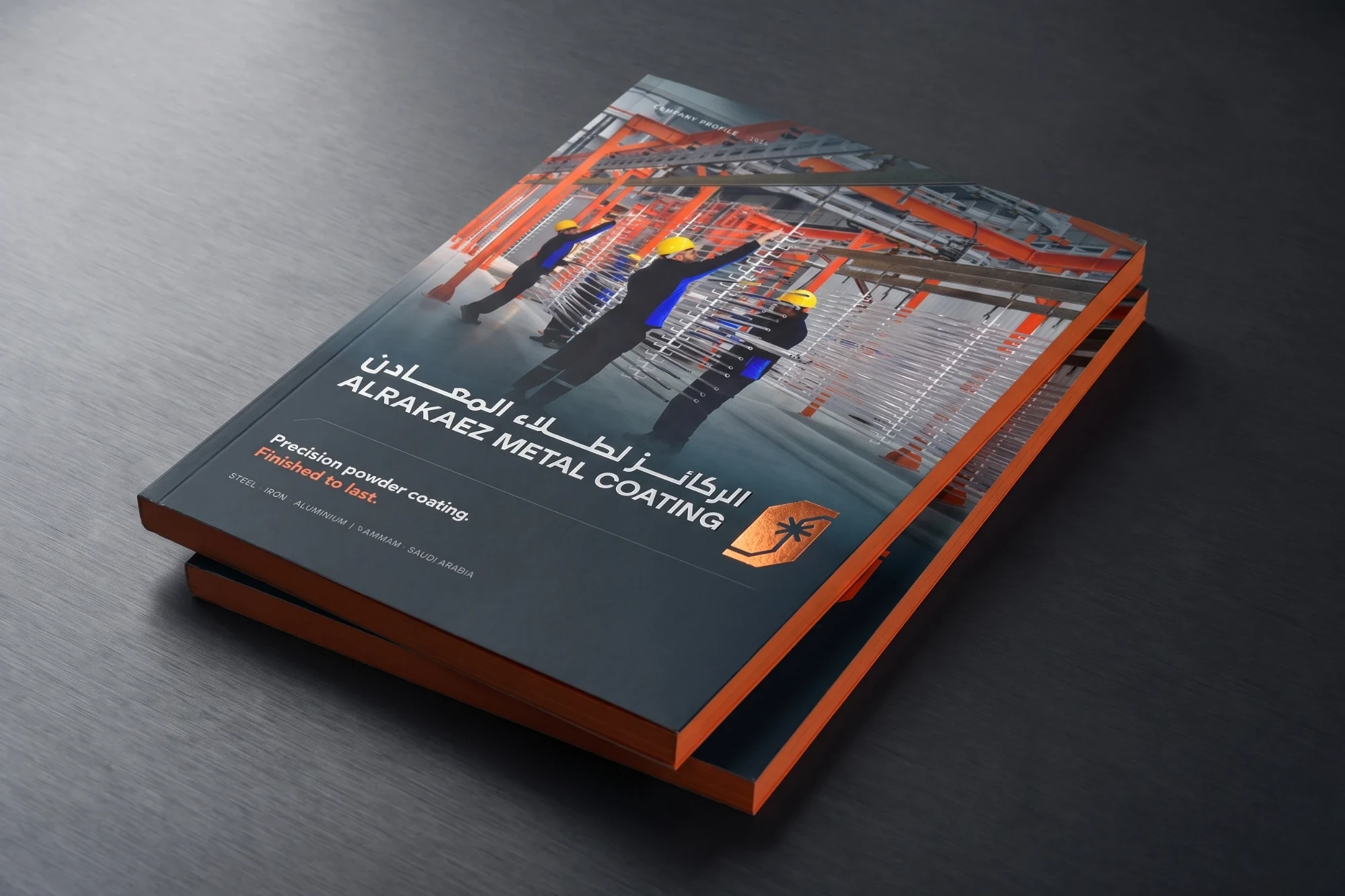 Mockup of two copies of the profile stacked on brushed metal. The cover shows workers in yellow hard hats at a powder-coating line, الركائز لطلاء المعادن and ALRAKAEZ METAL COATING, Precision powder coating. Finished to last., and the logo in copper foil.