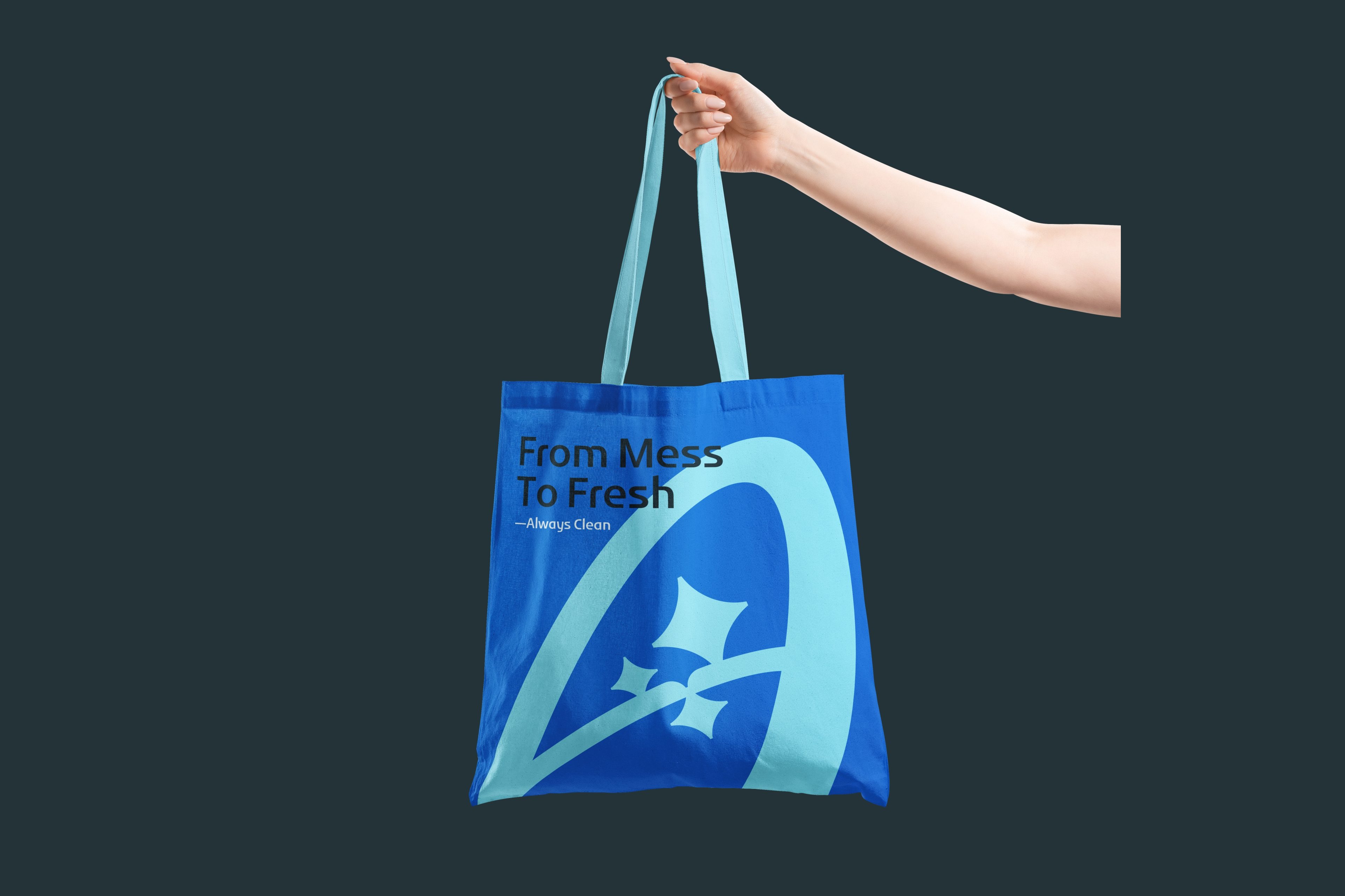 A hand holding a blue Always Clean tote bag printed with From Mess To Fresh and a large light blue monogram, on a dark background.
