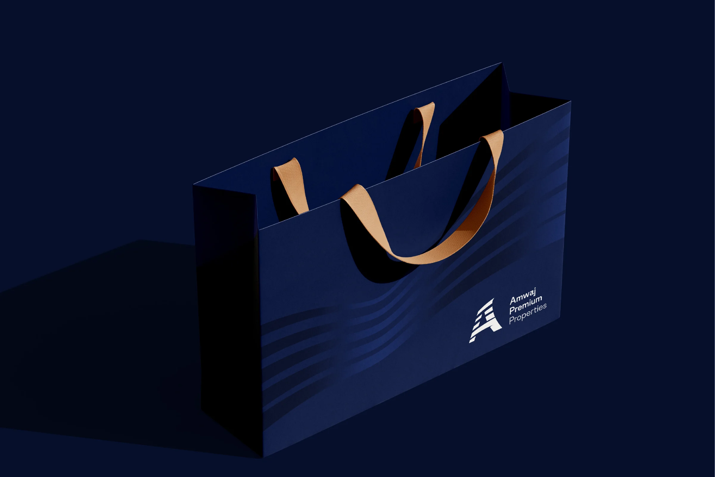 A navy paper bag with sand-coloured ribbon handles, tone-on-tone wave bands and the white logo, on a dark navy background.