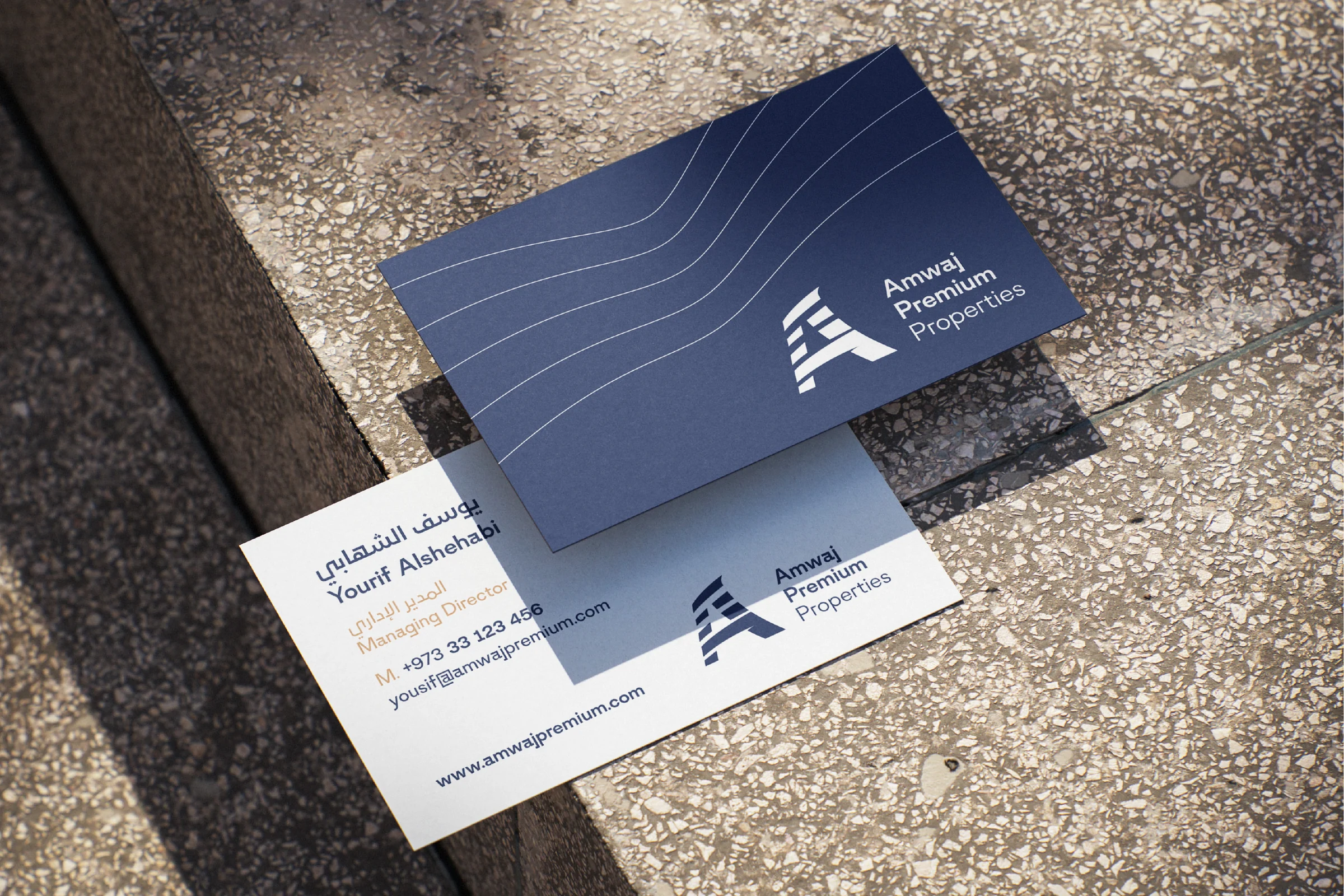 Two business cards on terrazzo steps: a navy back with fine white wave lines and the logo, and a white front with a name and title in Arabic and English, contact details and the logo.