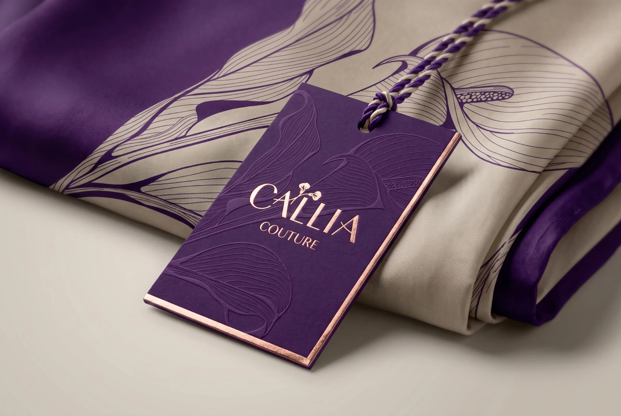 A plum hang tag with blind-embossed lilies, the logo in rose-gold foil and a rose-gold edge, tied with a plum and cream cord to a folded cream scarf printed with plum lilies.