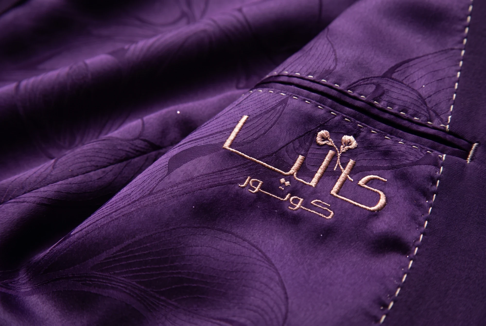 The Arabic logo embroidered in rose-gold thread on a plum jacket lining woven with lilies, beside a pocket.