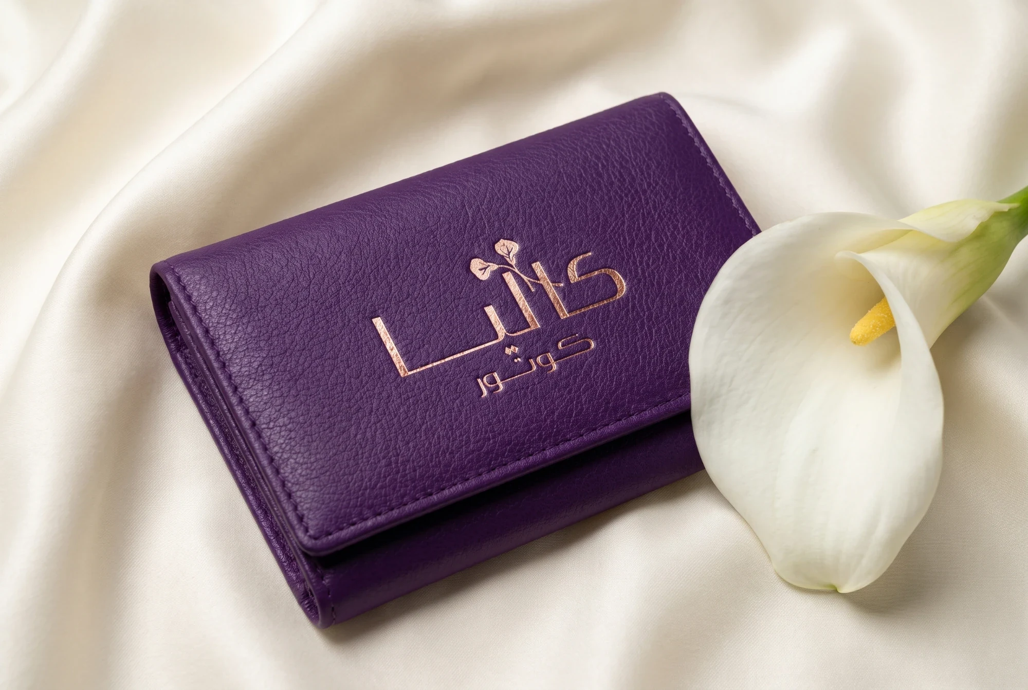 A plum leather wallet foiled with the Arabic logo in rose gold, on cream satin beside a white calla lily.