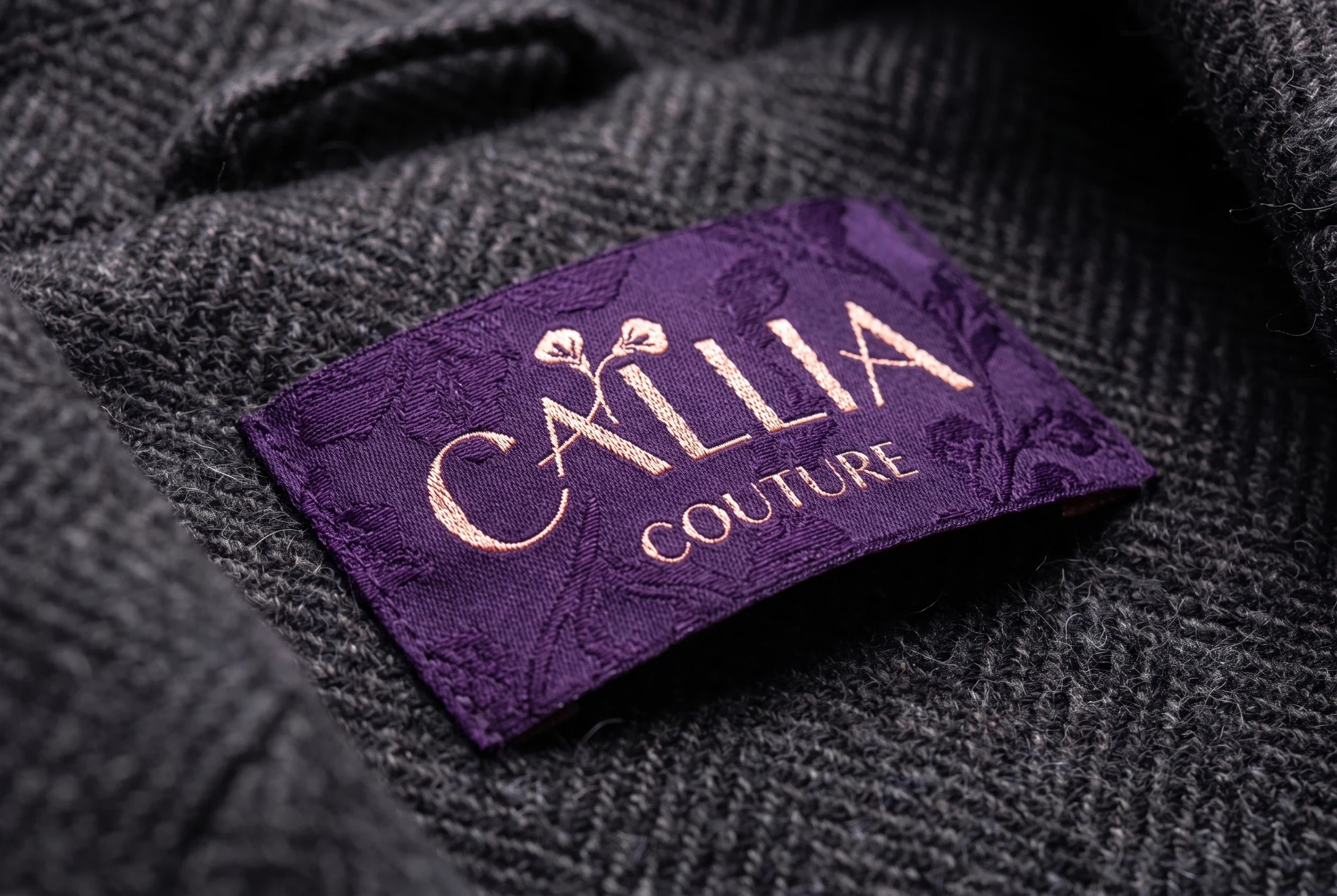 A plum woven label with a tone-on-tone lily jacquard and the logo in rose-gold thread, on a dark herringbone coat.