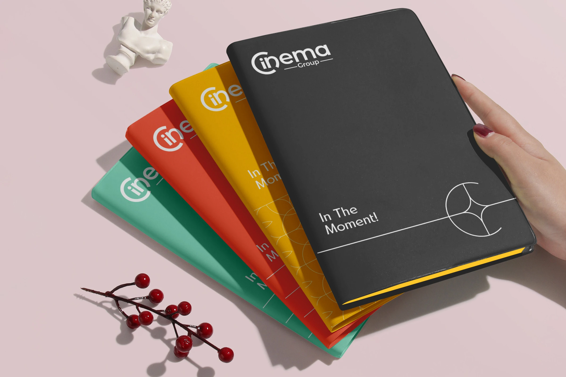 A hand fanning notebooks in black, yellow, coral and green, each with the logo, the black one with In The Moment! and a hairline figure.