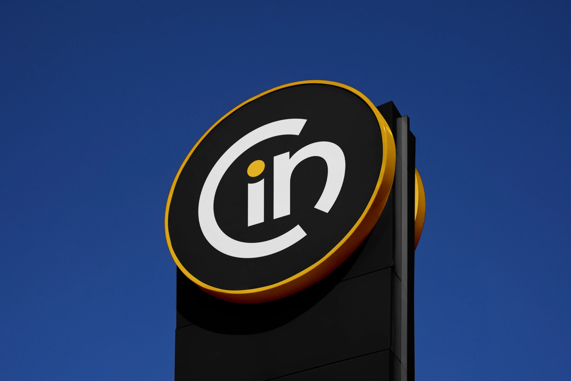 A round pylon sign with the symbol in white on black, edged in yellow, against a deep blue sky.