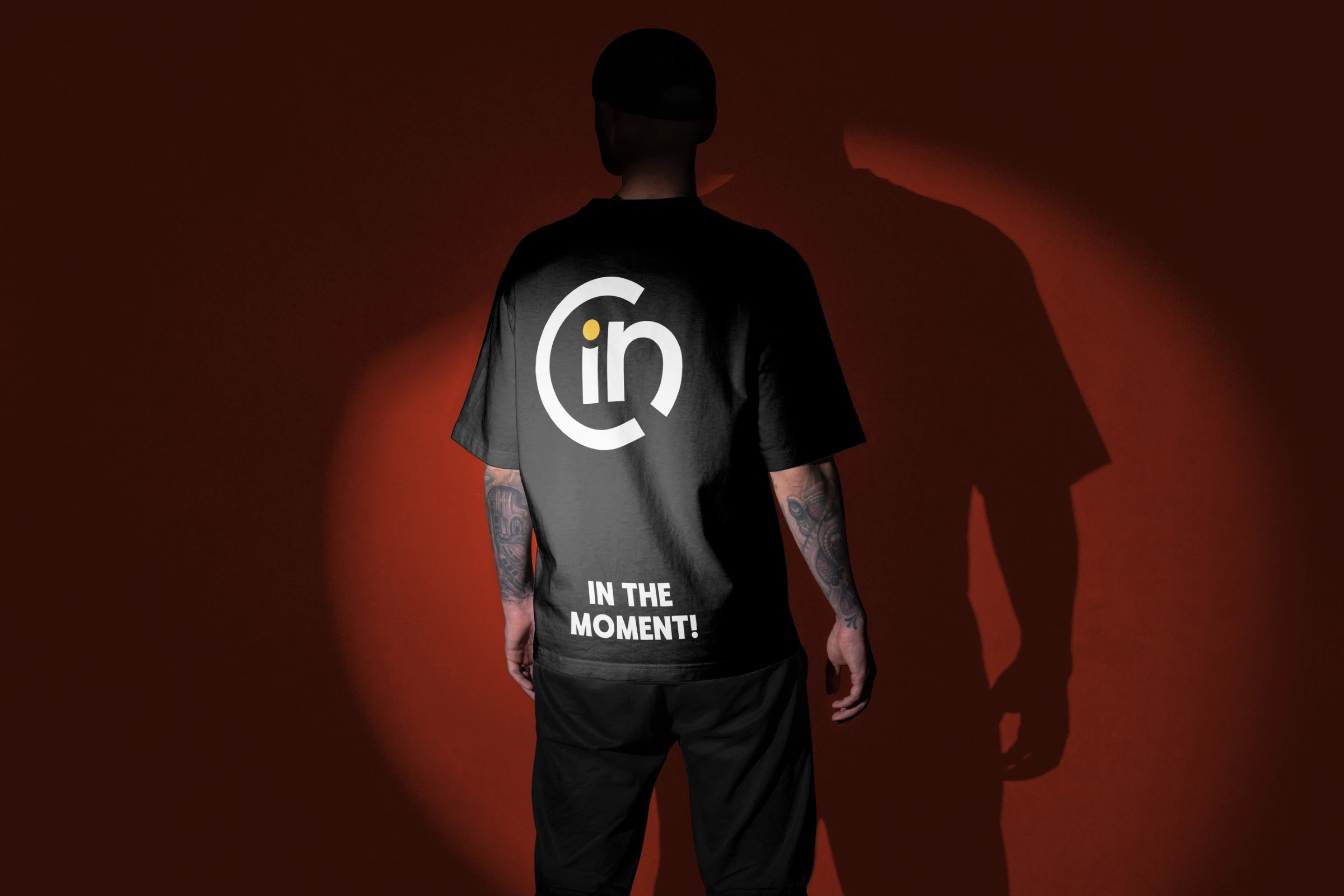 A man seen from behind in a black T-shirt with the symbol and IN THE MOMENT! printed on the back, in a red spotlight.