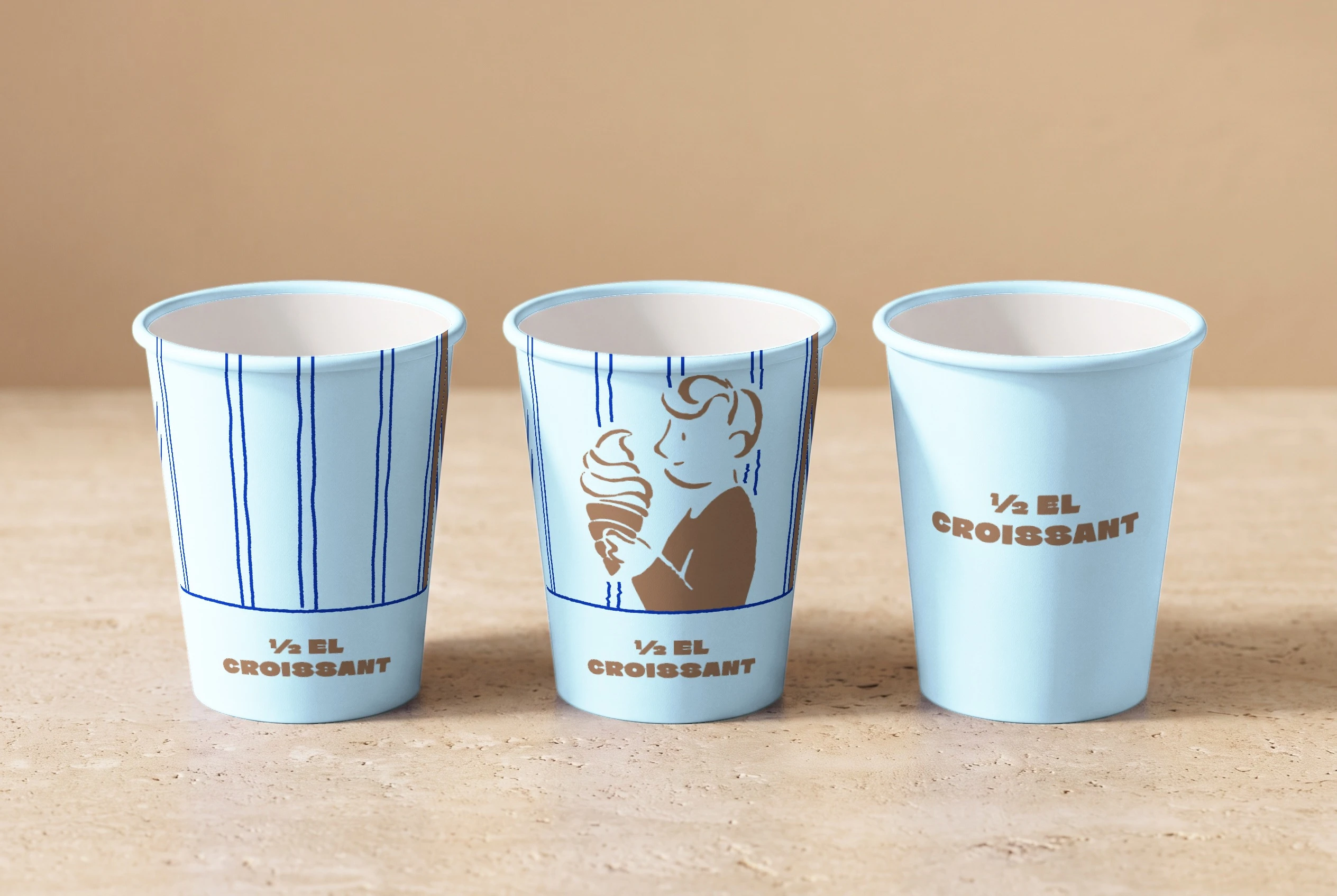 Three baby-blue paper cups on a travertine counter: one with navy stripes, one with the boy and his croissant cone under the stripes, and a plain one with the wordmark; the first two carry ½ EL CROISSANT on a band at the bottom.