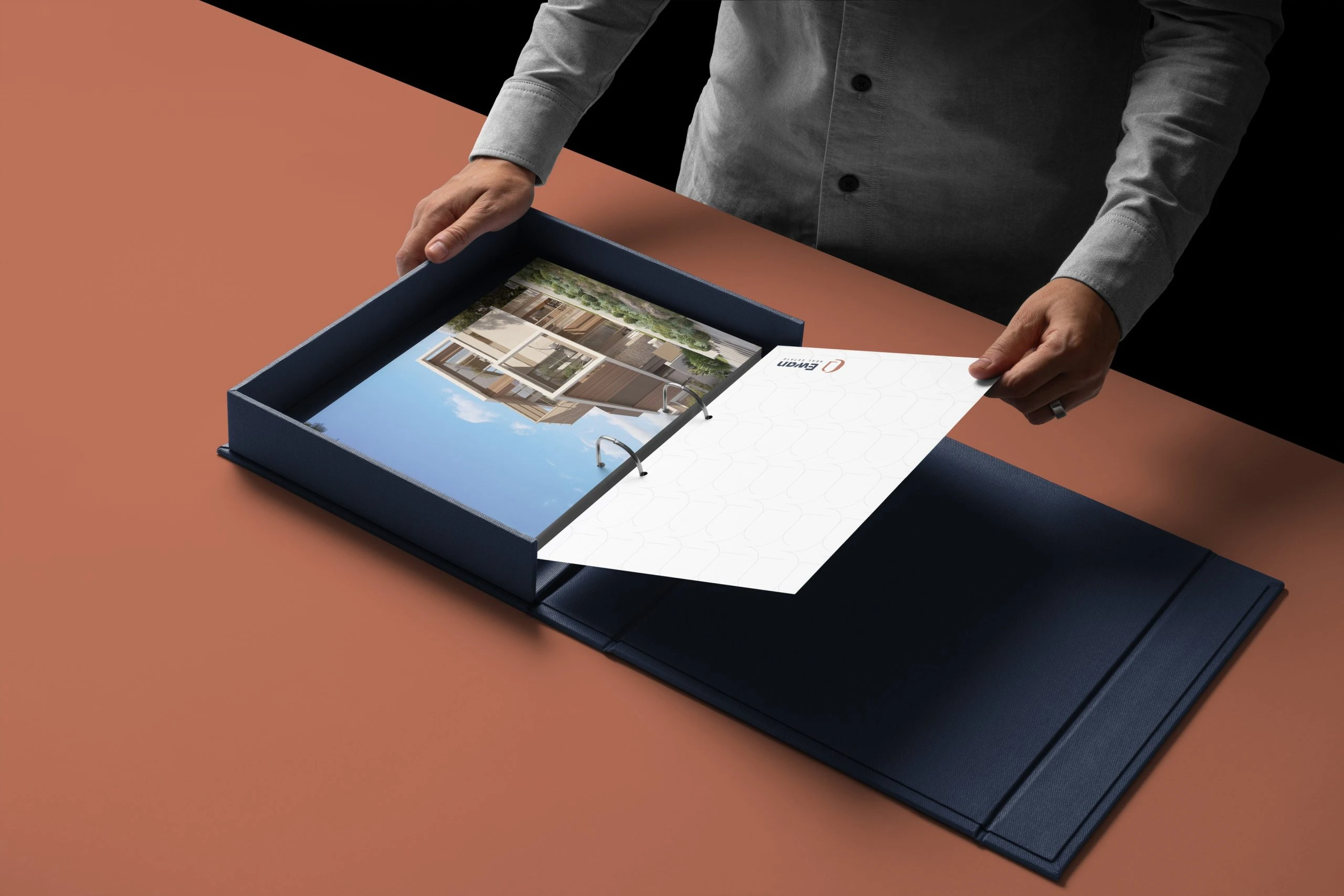 A navy presentation box open on a terracotta table, hands in grey sleeves turning a white binder page with the Ewan Real Estate logo over a photo of a modern building.