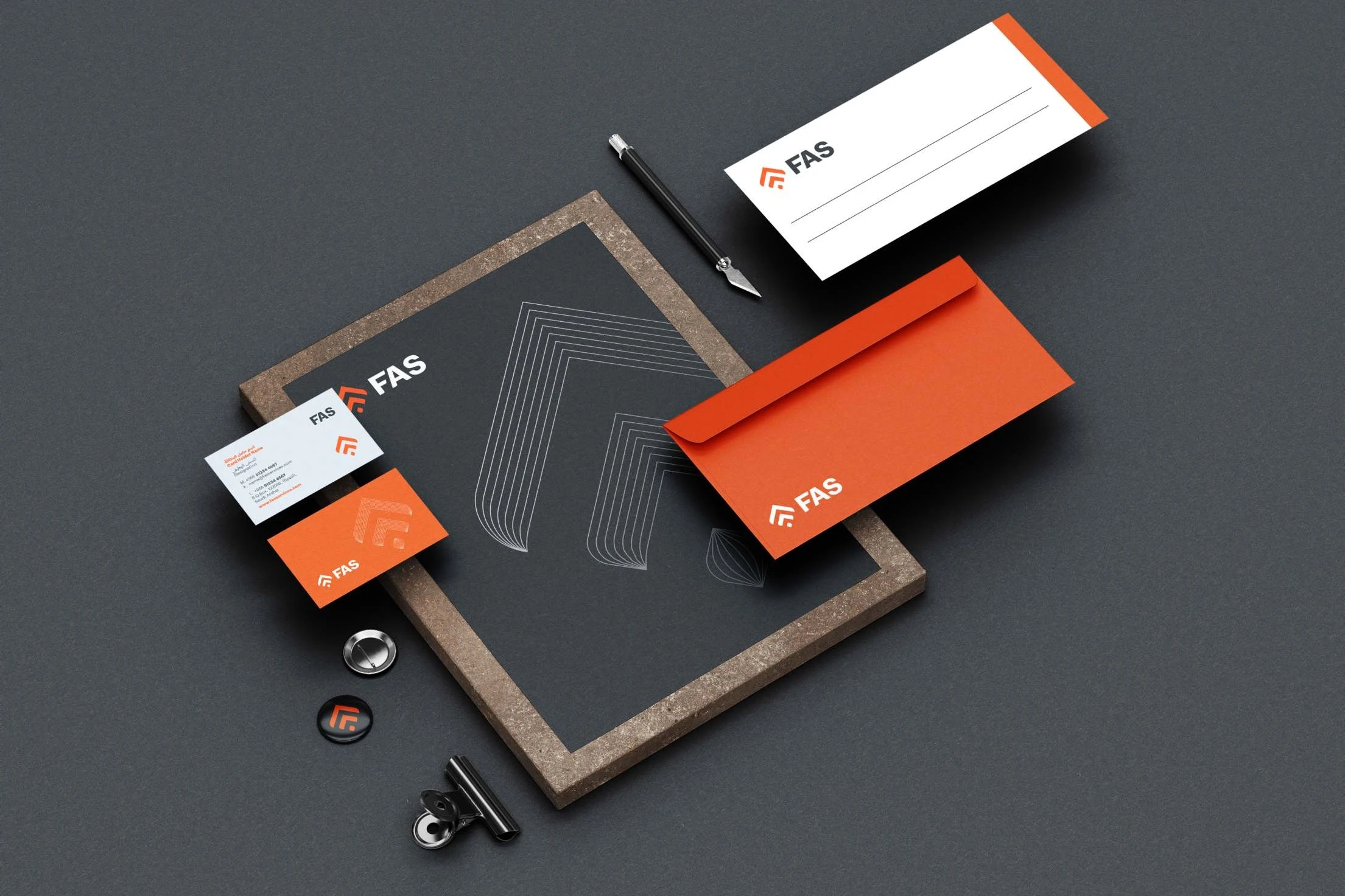 Stationery on dark grey: a slate sheet with the logo and the line-drawn mark, white and orange business cards, an orange envelope, a lined white slip, a craft knife and pin badges.