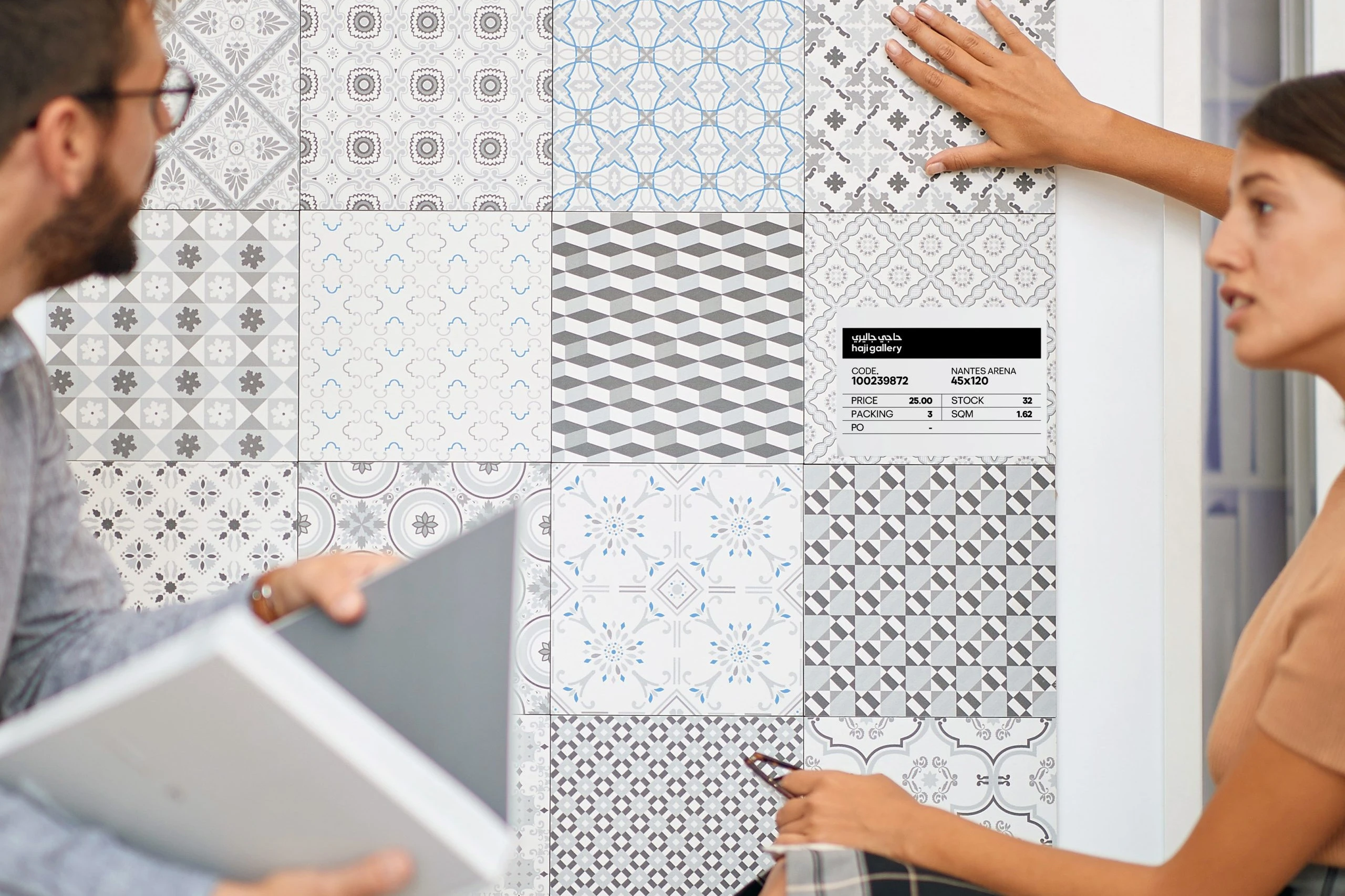Two people at a wall of patterned grey, white and blue tiles, one touching a tile, beside a Haji Gallery price tag with the tile's code, size, price and stock.
