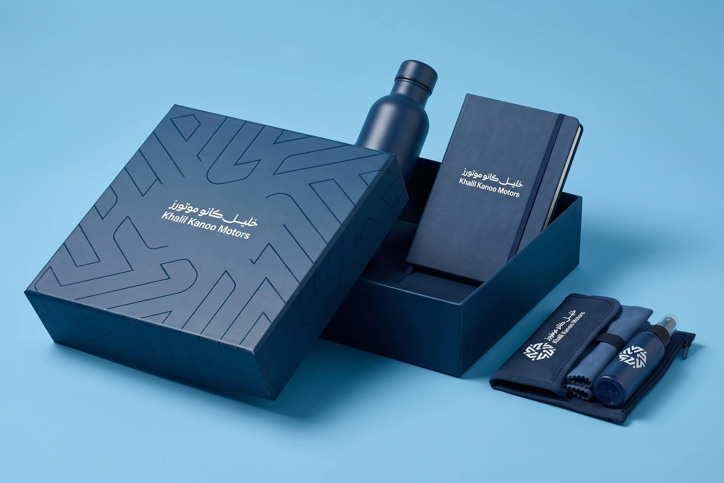 A navy gift box printed with the mark’s strokes and the logo, with a navy bottle, a notebook and a car-care pouch, on sky blue.
