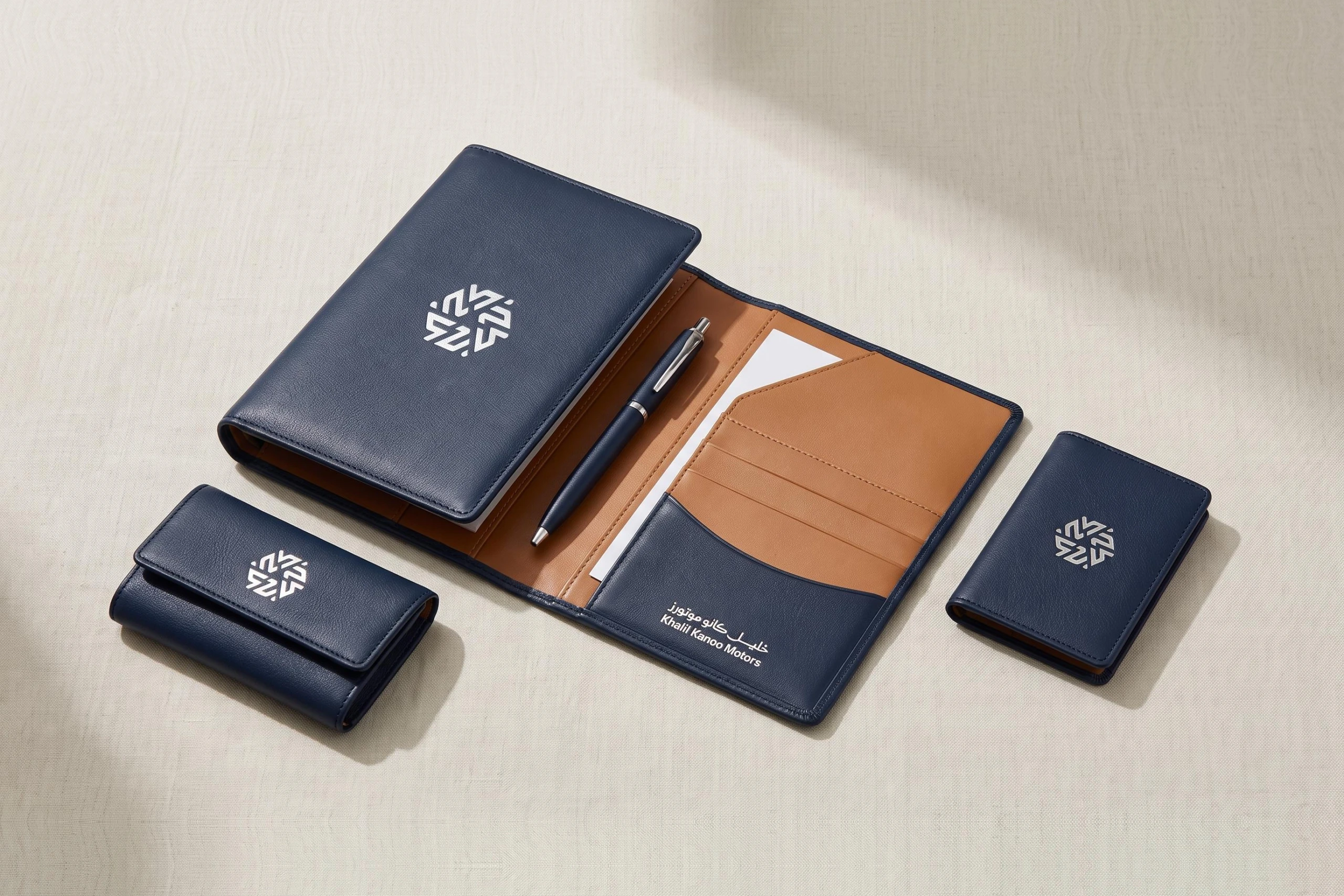 A navy leather folio with a tan lining, a key case and a card case, each with the mark in silver, and the logo on the folio’s pocket.