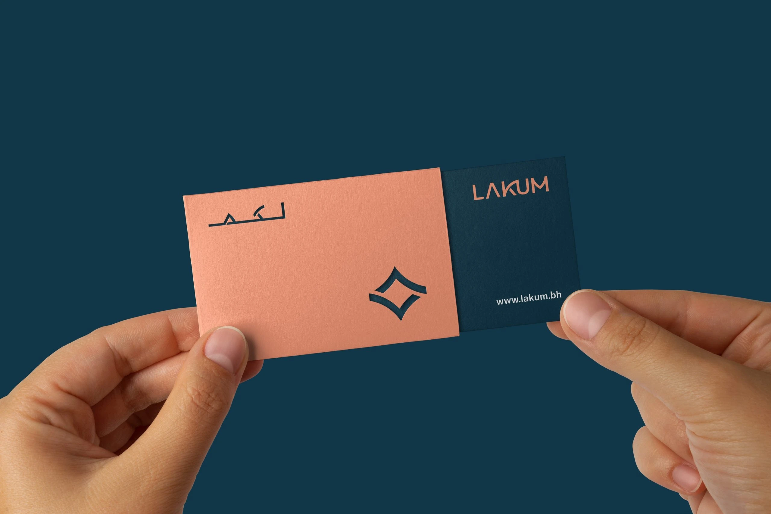 Lakum business cards held in two hands: a coral card with لكم and the emblem cut out, over a deep blue card with LAKUM in coral and www.lakum.bh.