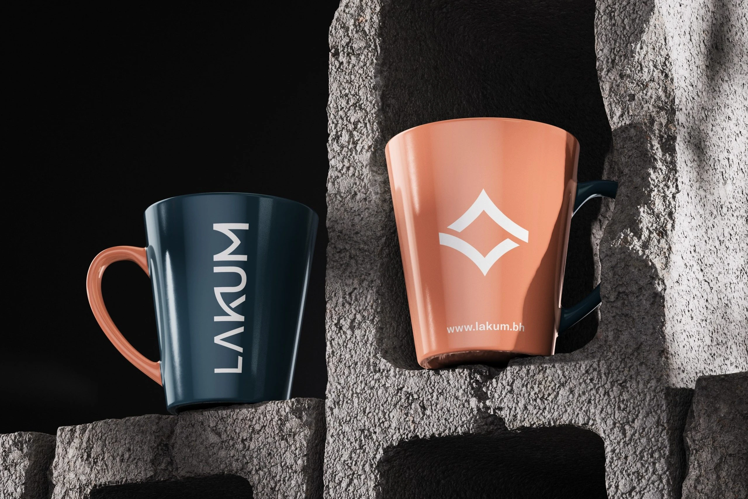 Two mugs on rough concrete blocks: a deep blue mug with LAKUM in white and a coral handle, and a coral mug with the white emblem and www.lakum.bh.