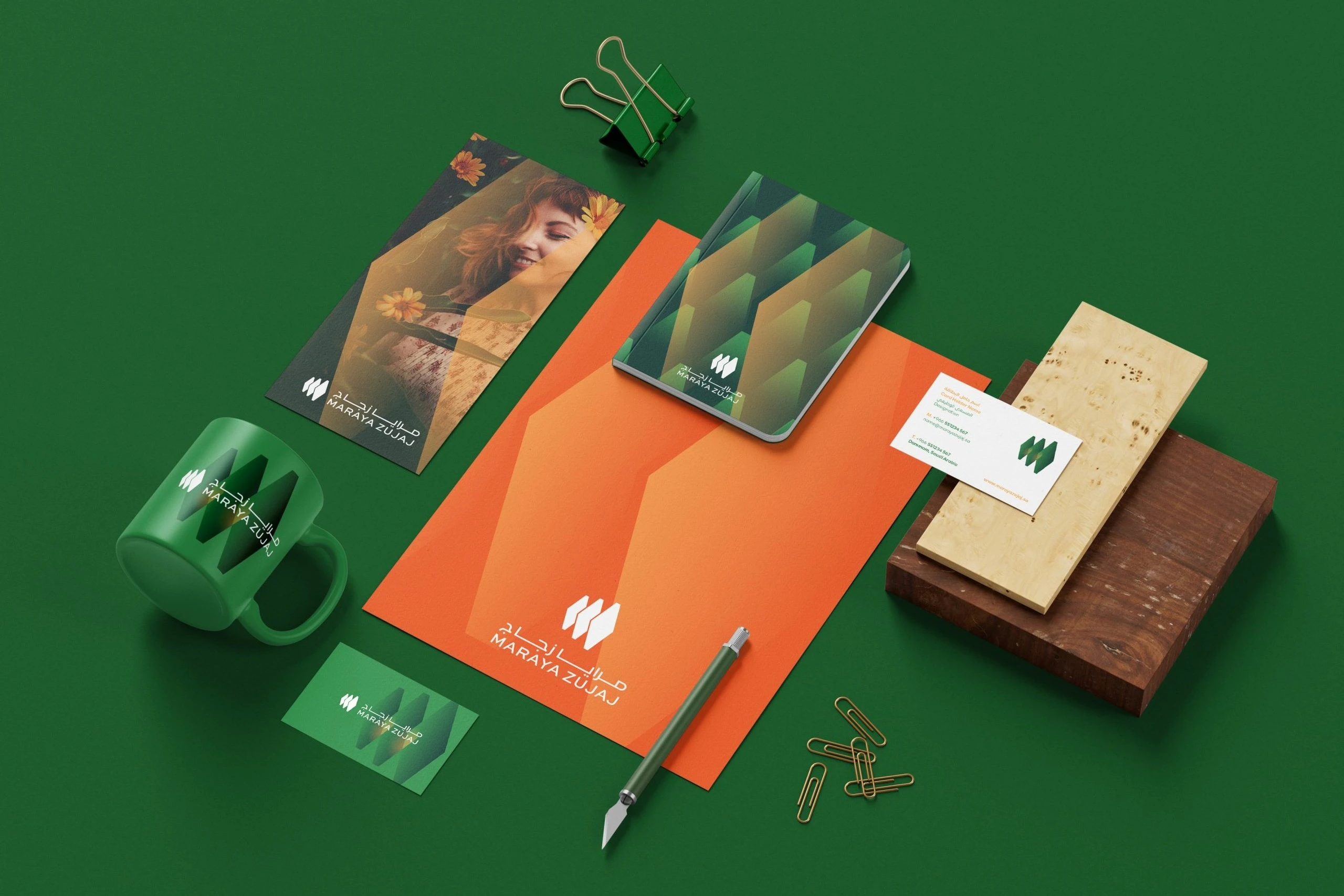 Stationery on green: an orange letterhead, a notebook in the hexagon pattern, a green mug, business cards, a flyer with a portrait photo, a craft knife and clips.