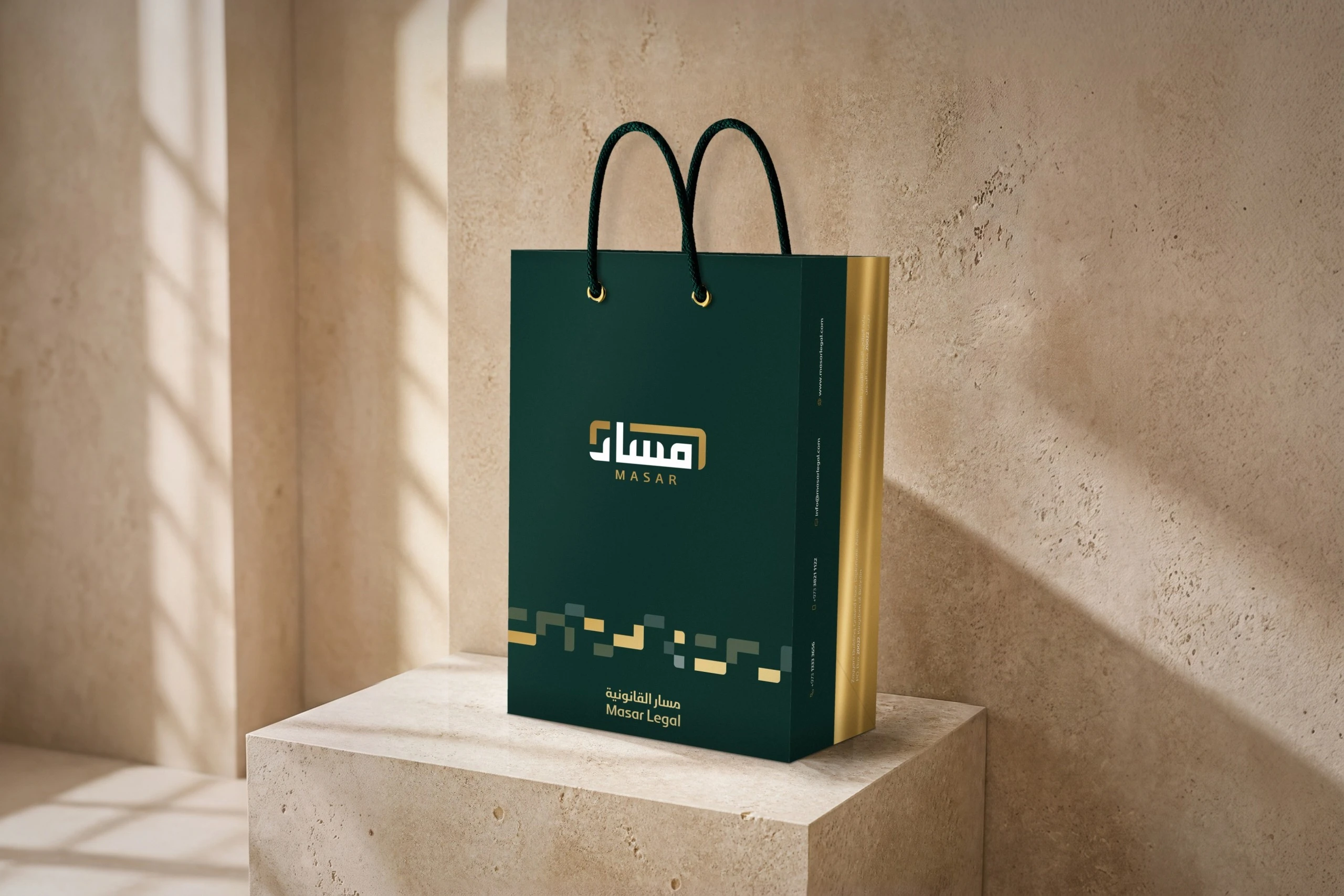 A deep green Masar paper bag with dark green rope handles and gold eyelets on a travertine plinth; the gold half of the side panel carries the address.