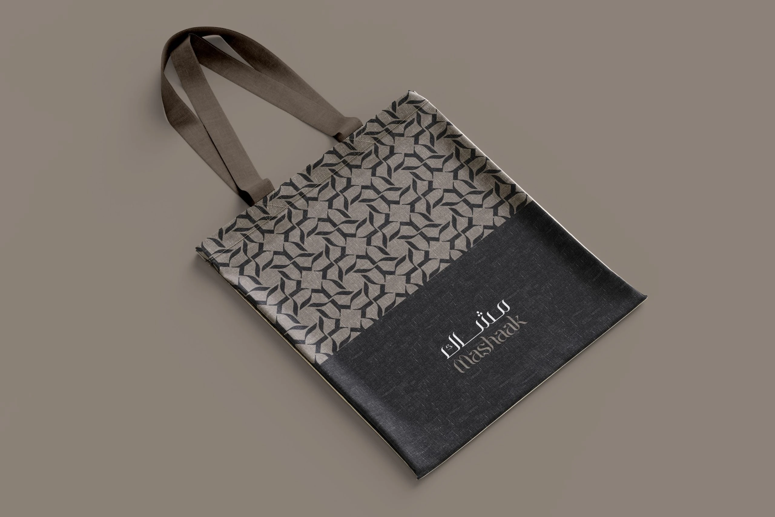 A Mashaak tote bag with a patterned top and a charcoal base carrying the bilingual logotype, on taupe.
