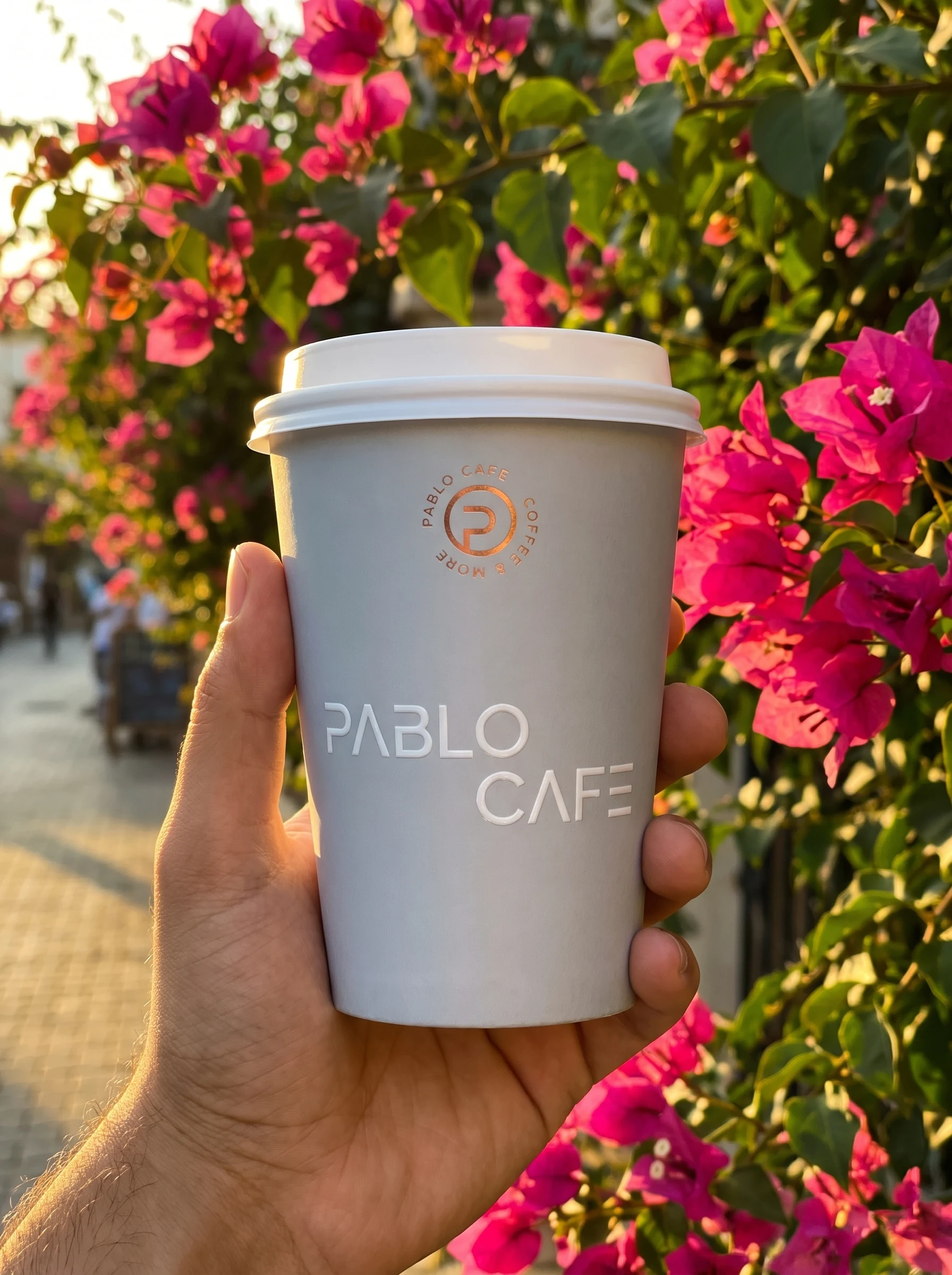A hand holding a pale grey cup against bougainvillea, with the roundel in copper foil and PABLO CAFE in white relief.