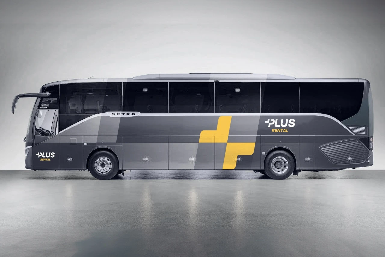 A grey bus in a studio, the Plus Rental logo at both ends of its side and a large yellow split plus across the middle.
