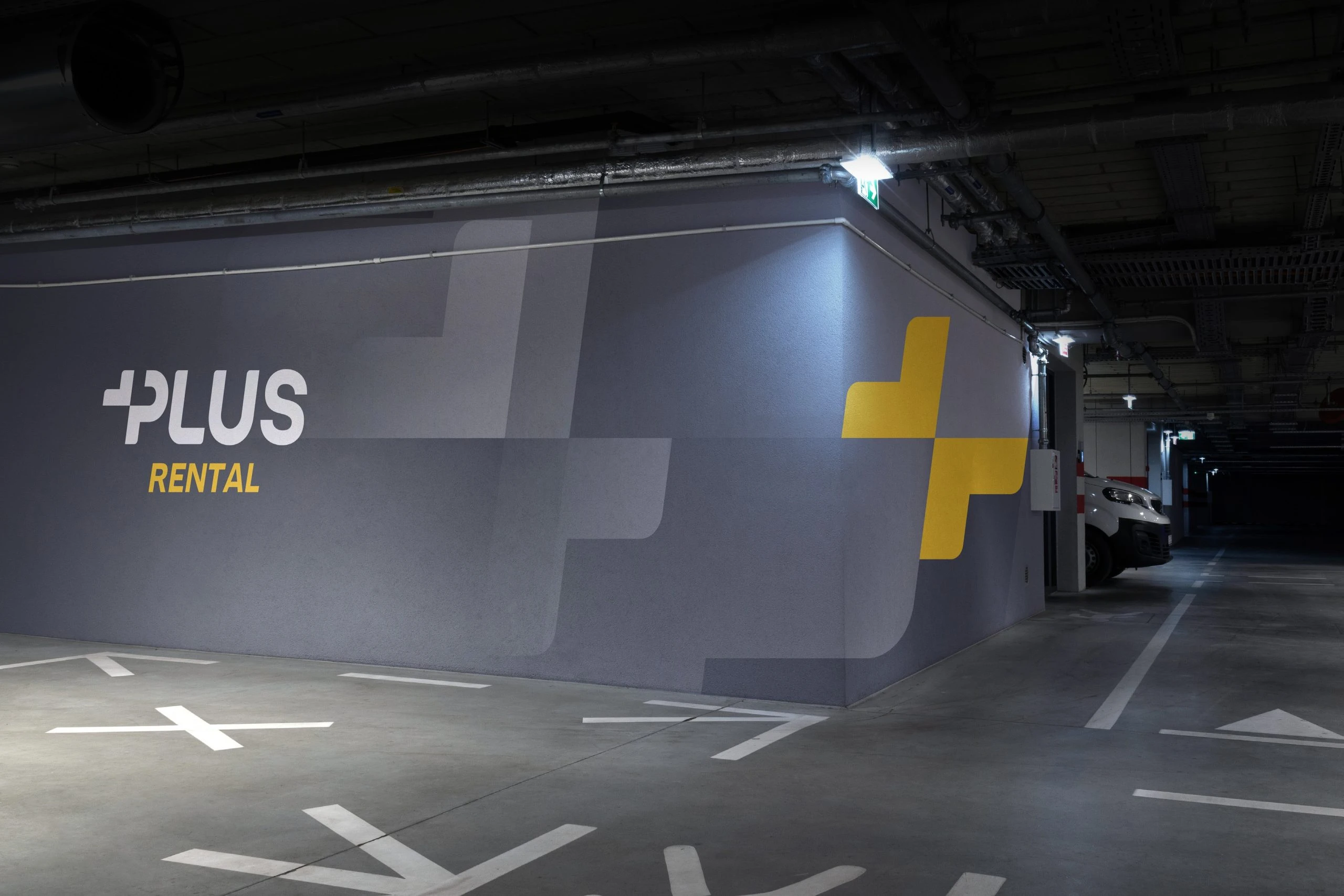 A slate wall in an underground car park with the Plus Rental logo in white and yellow and a large split yellow plus wrapping the corner, beside a parked white van.