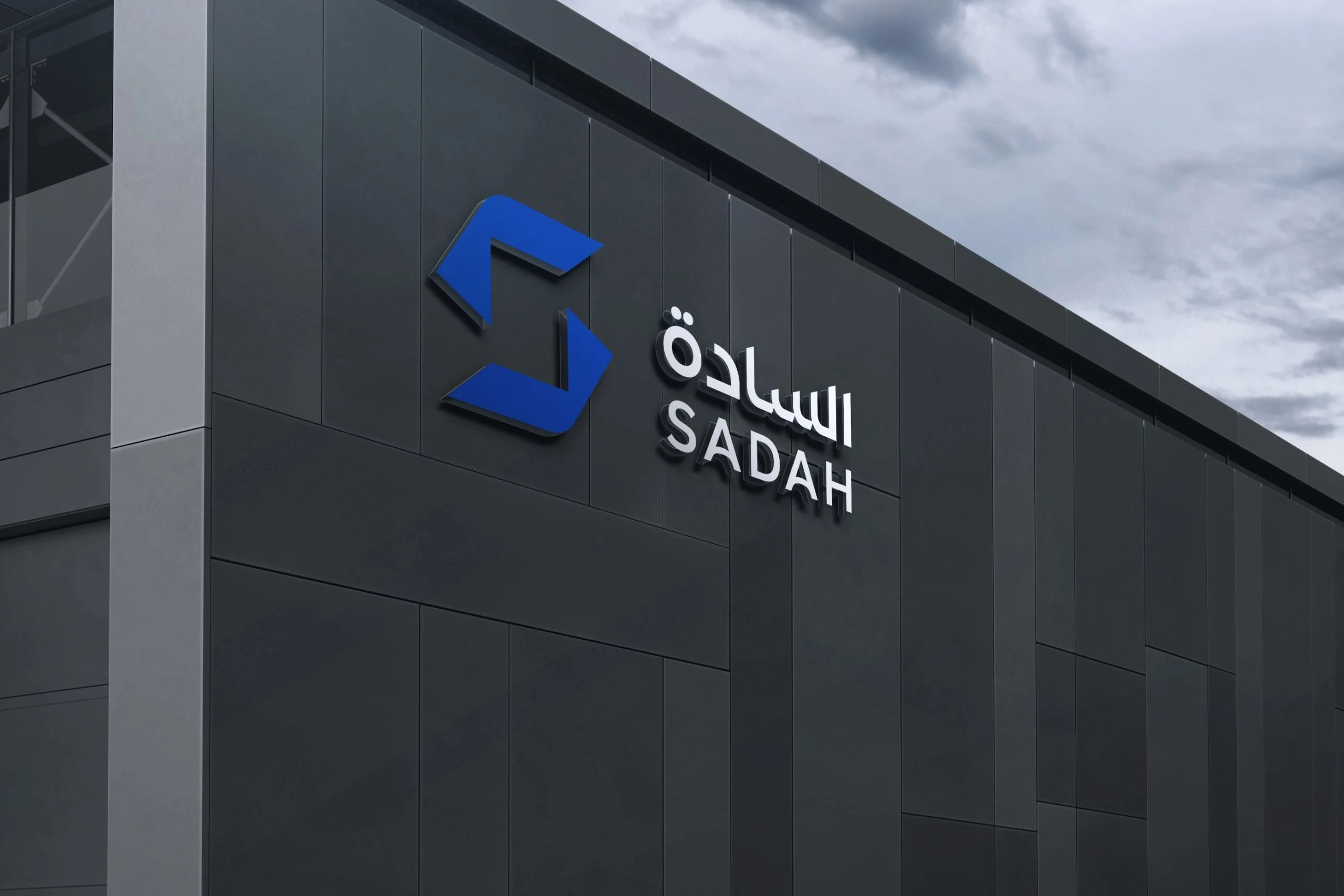 The Sadah logo in raised letters, the blue S and white names, on a dark ribbed building under a cloudy sky.