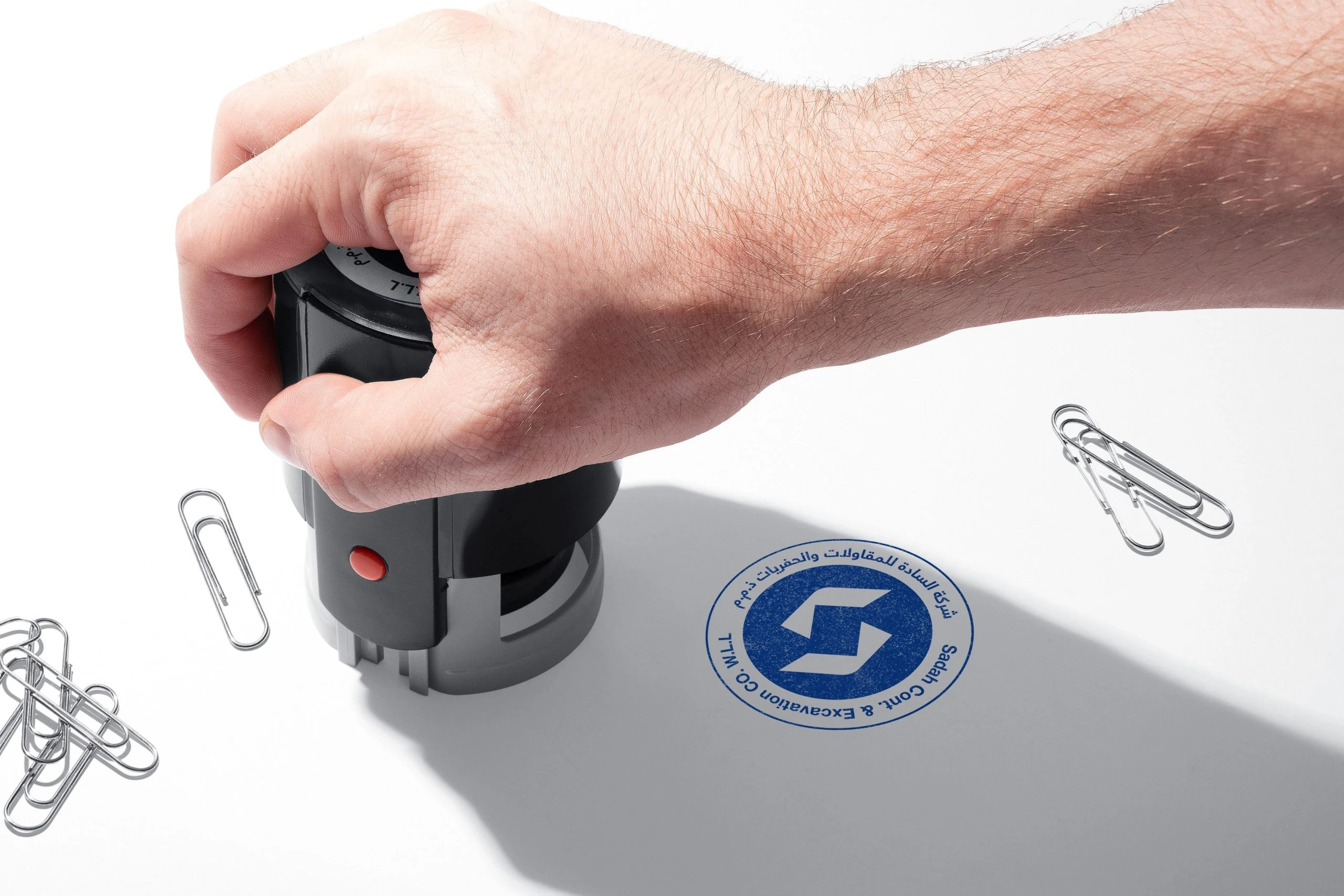 A hand pressing a self-inking stamp beside its blue round impression with the S symbol, among paper clips.