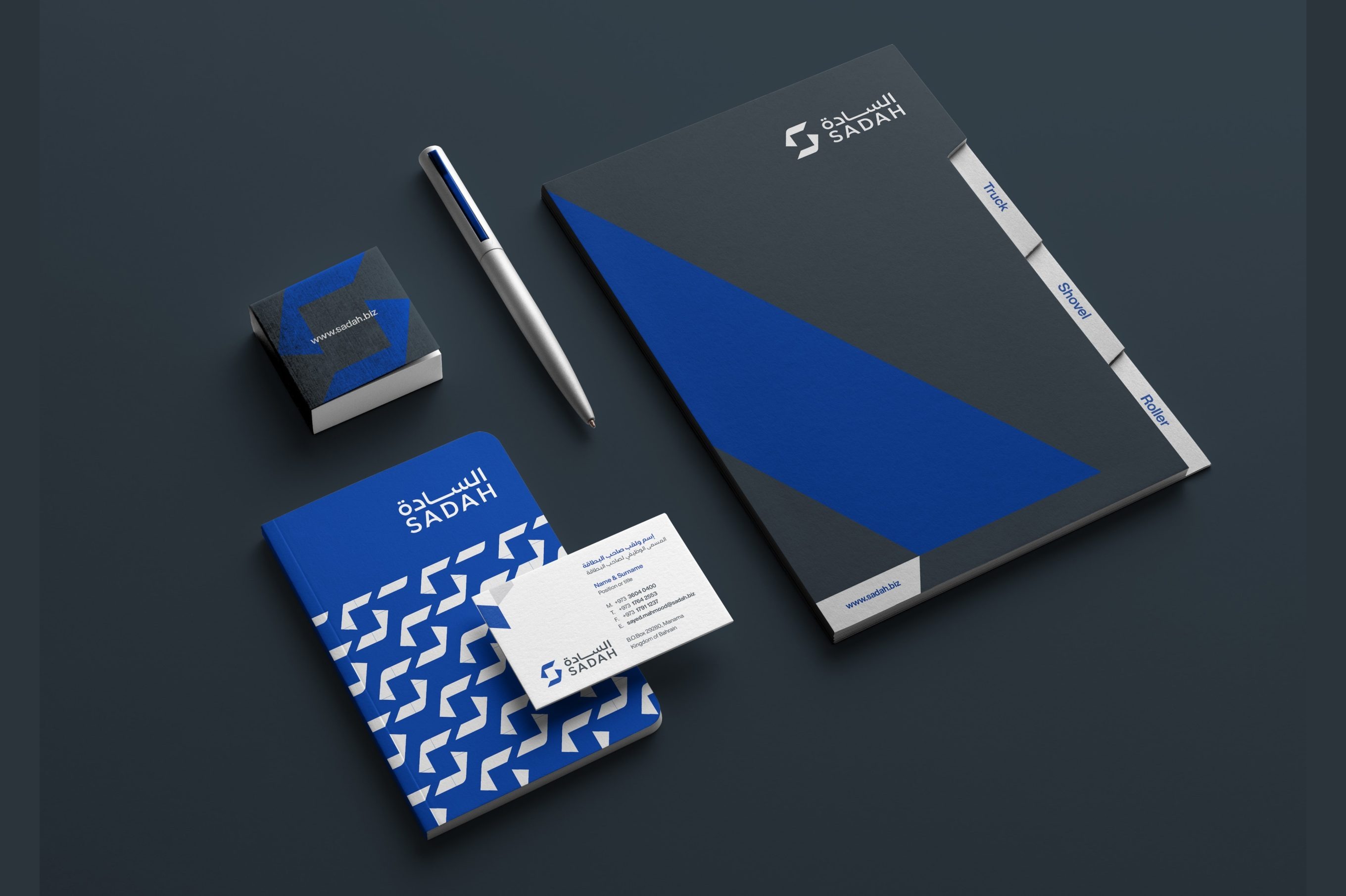 Sadah stationery on dark slate: a folder with a blue diagonal and index tabs, a notebook covered in the blue S pattern, a business card, a pen and a sticky-note block.