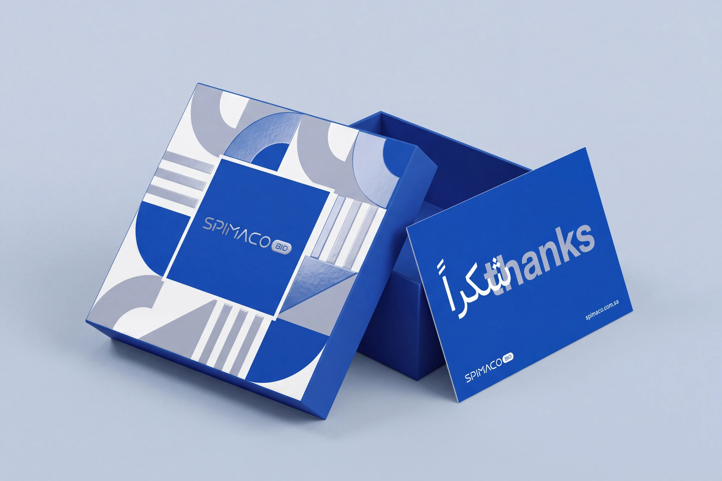 An open blue gift box, its lid covered in the pattern with the wordmark in a blue square, beside a blue card reading شكراً and thanks, with the wordmark and spimaco.com.sa.
