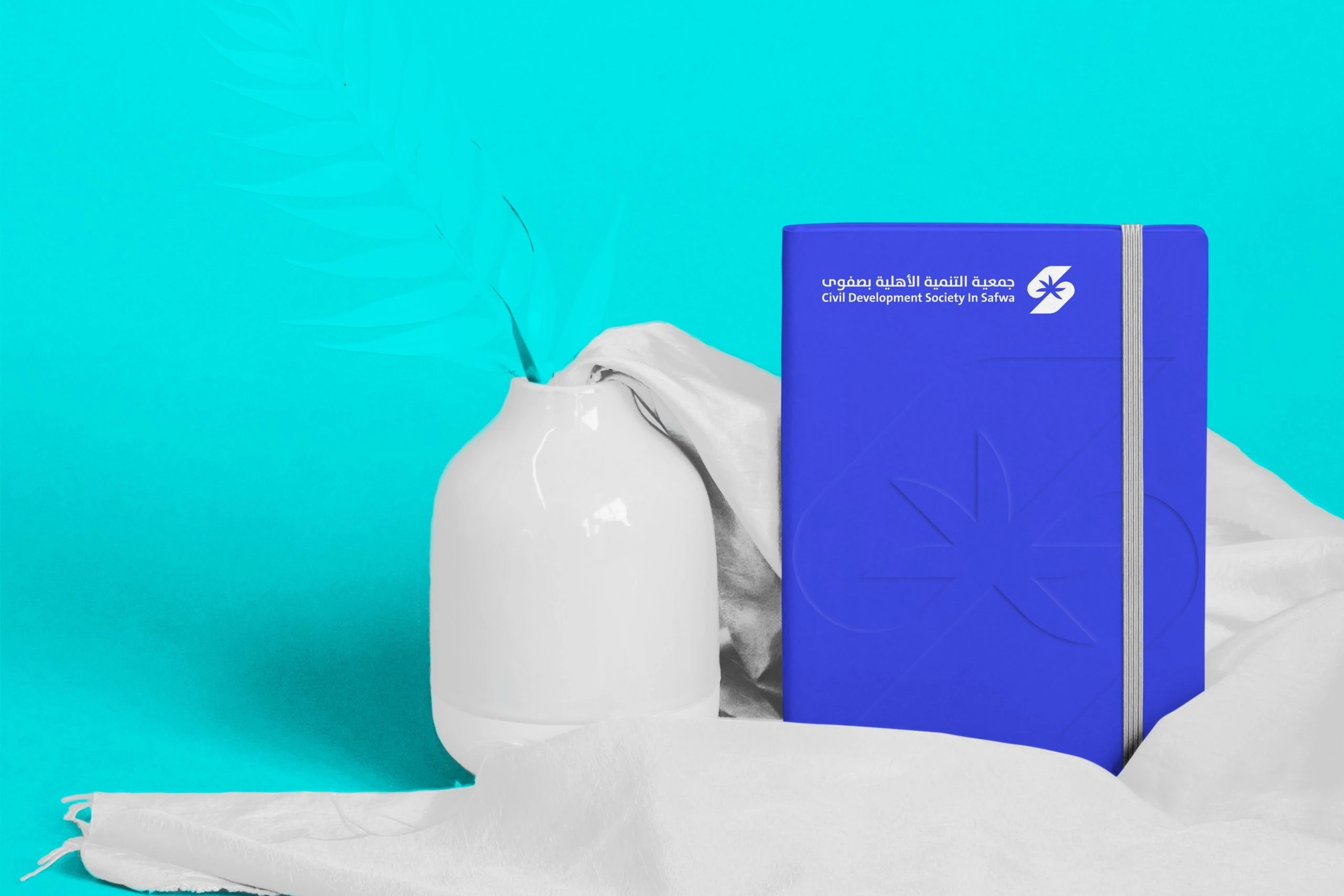 A royal blue notebook with the logo, standing beside a white vase on white cloth against turquoise.