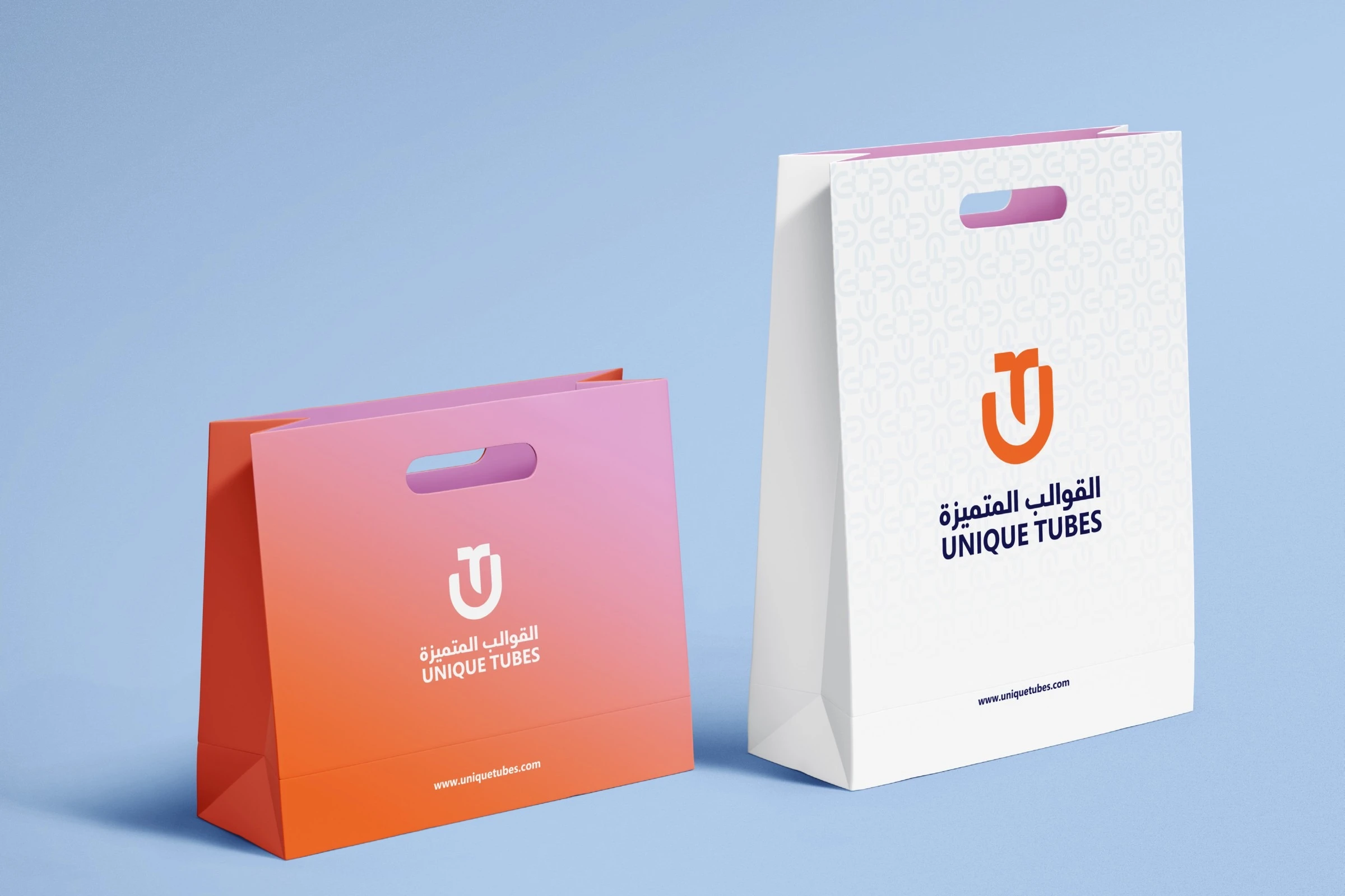 Two paper bags on pale blue: a pink-to-orange bag with the logo in white, and a white bag with a faint monogram pattern and the logo in orange and dark blue.