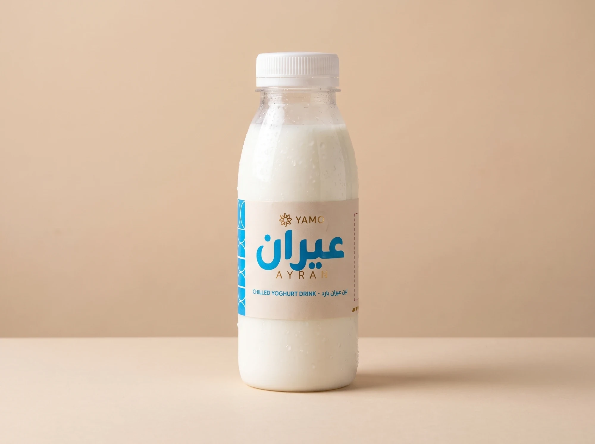 A bottle of ayran with a cream label: عيران in blue, AYRAN in gold and CHILLED YOGHURT DRINK.