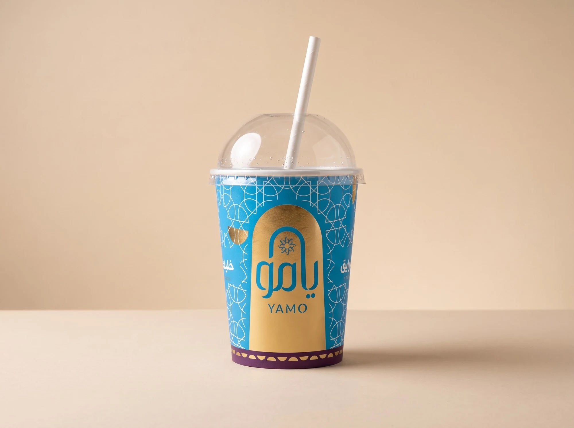A blue cold cup with a domed lid and a straw, the lattice in white and the logo in blue on a gold arch, with a plum band of half-moons at its foot.