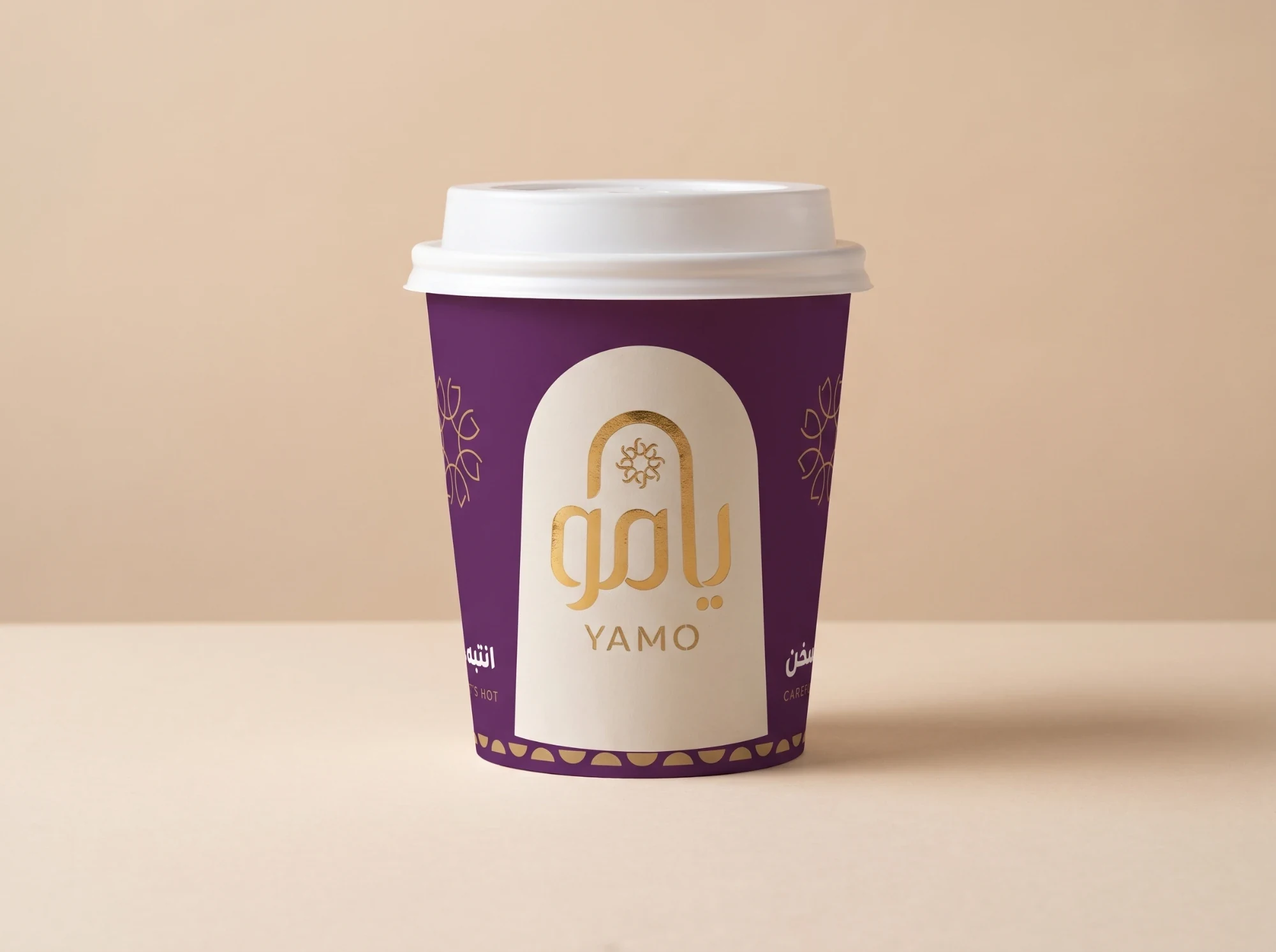 A plum paper cup with the logo in gold foil inside a cream arch and a row of gold half-moons at its foot.