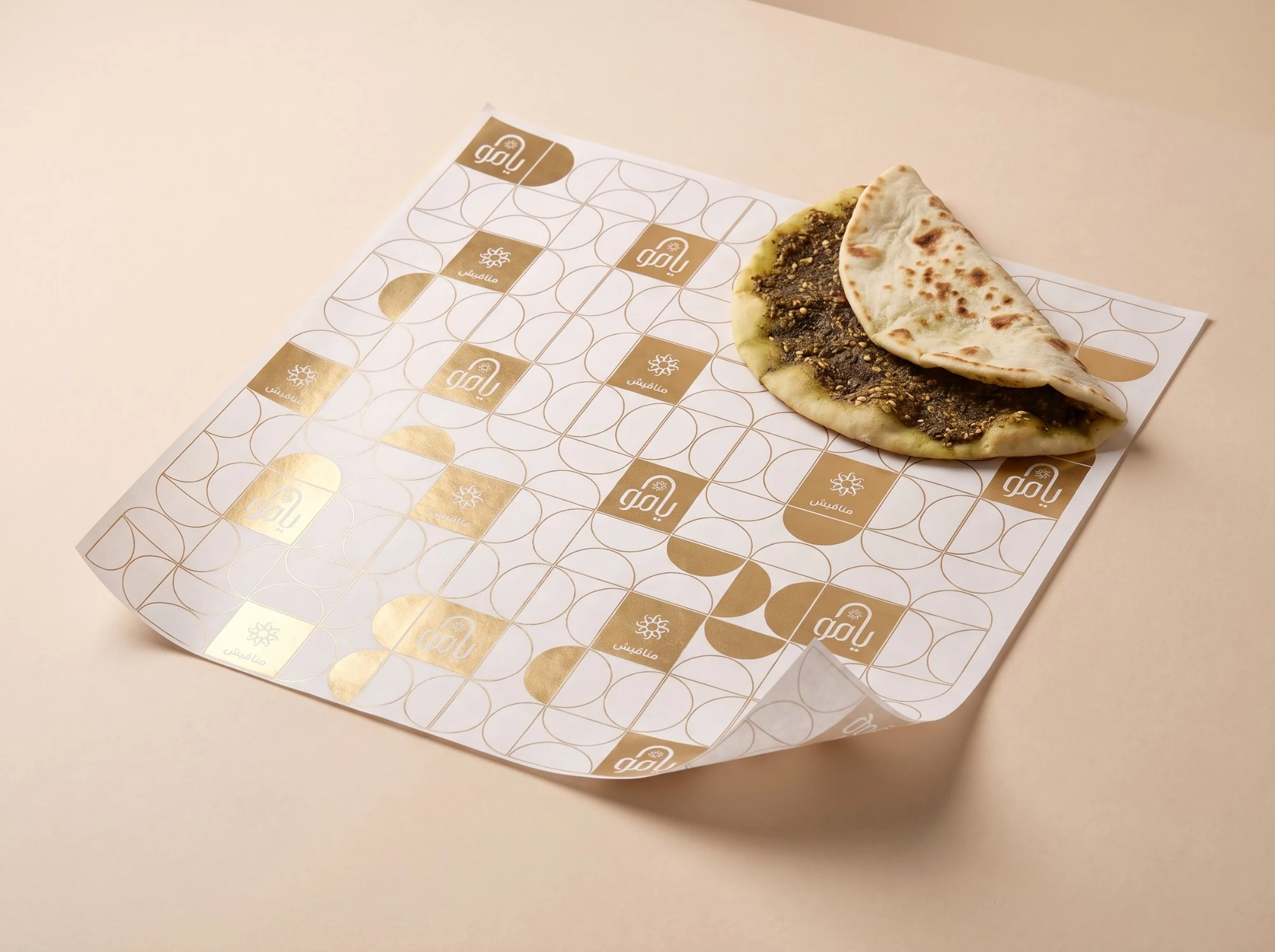 White paper printed with the half-moon grid in gold, with gold tiles carrying يامو and مناقيش, under a folded zaatar manousheh.