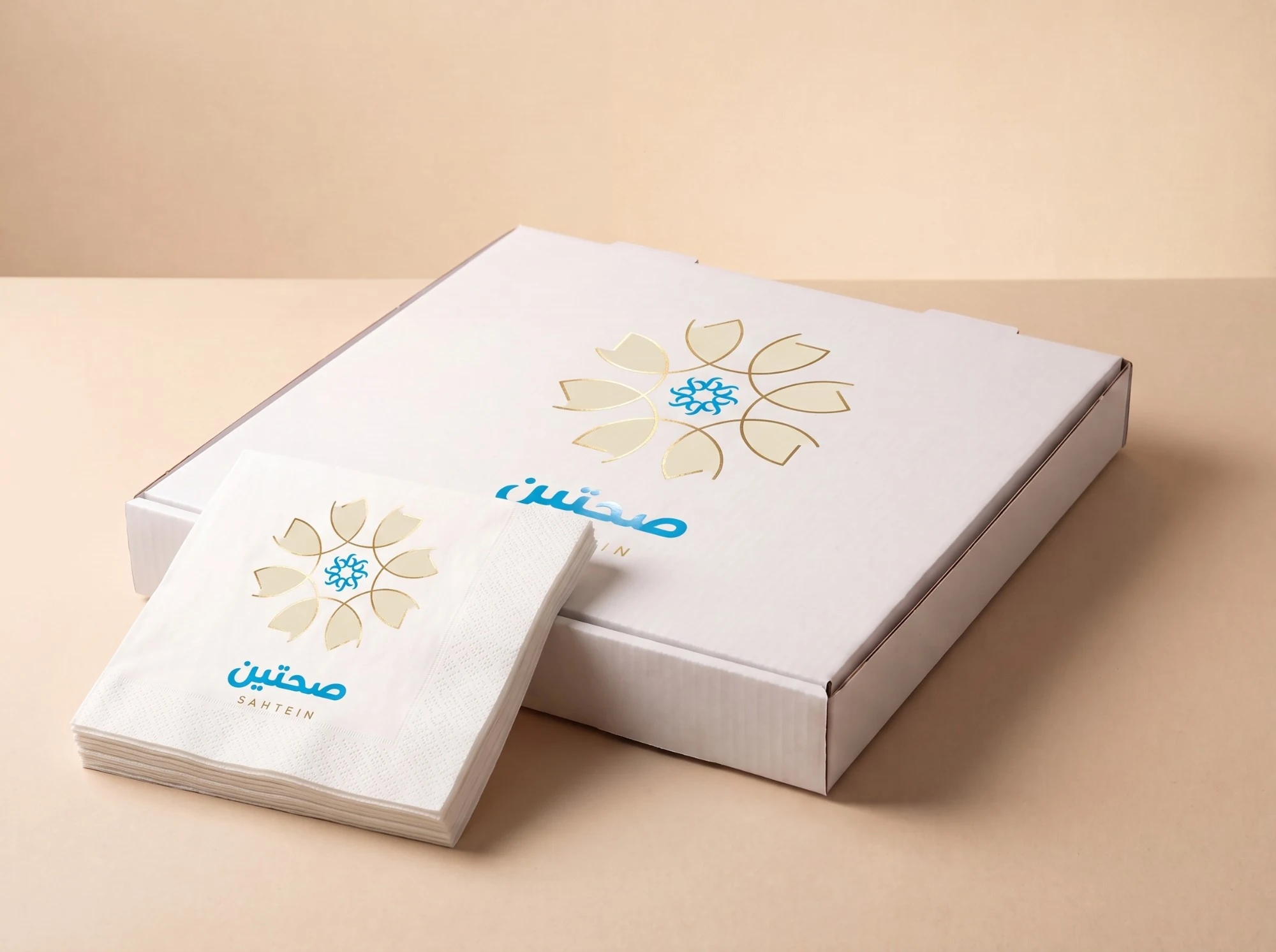 A stack of napkins and a white box, each with the rosette enlarged in gold and صحتين, SAHTEIN, in blue.