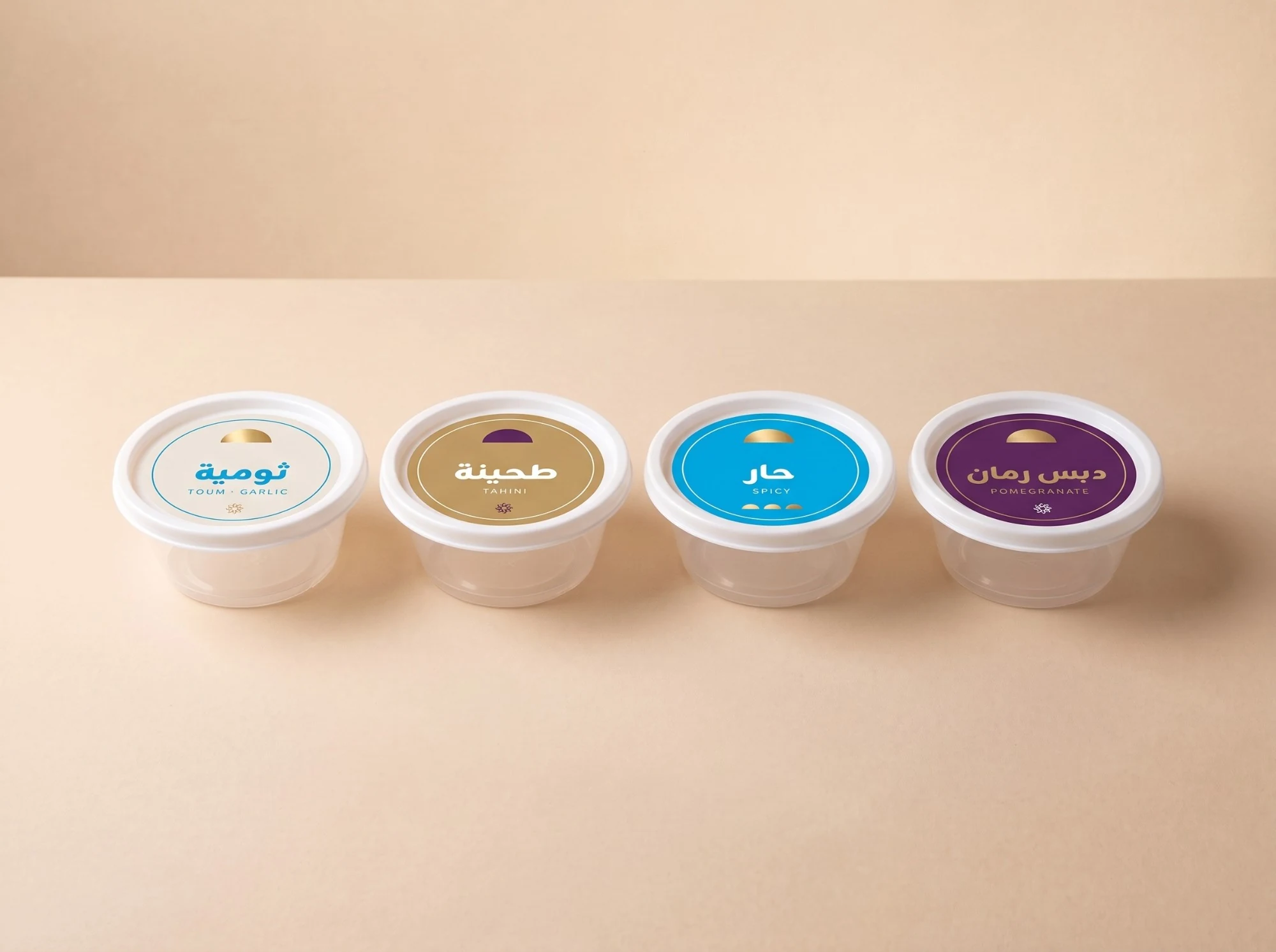 Four sauce pots with round lid labels in cream, gold, blue and plum: toum, tahini, spicy and pomegranate.