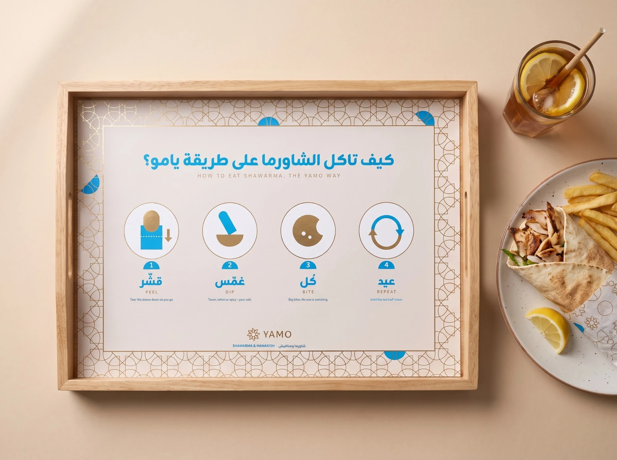 A wooden tray with a cream liner, كيف تاكل الشاورما على طريقة يامو؟, HOW TO EAT SHAWARMA, THE YAMO WAY, and four steps in blue and gold: peel, dip, bite and repeat, beside a shawarma plate and iced tea.